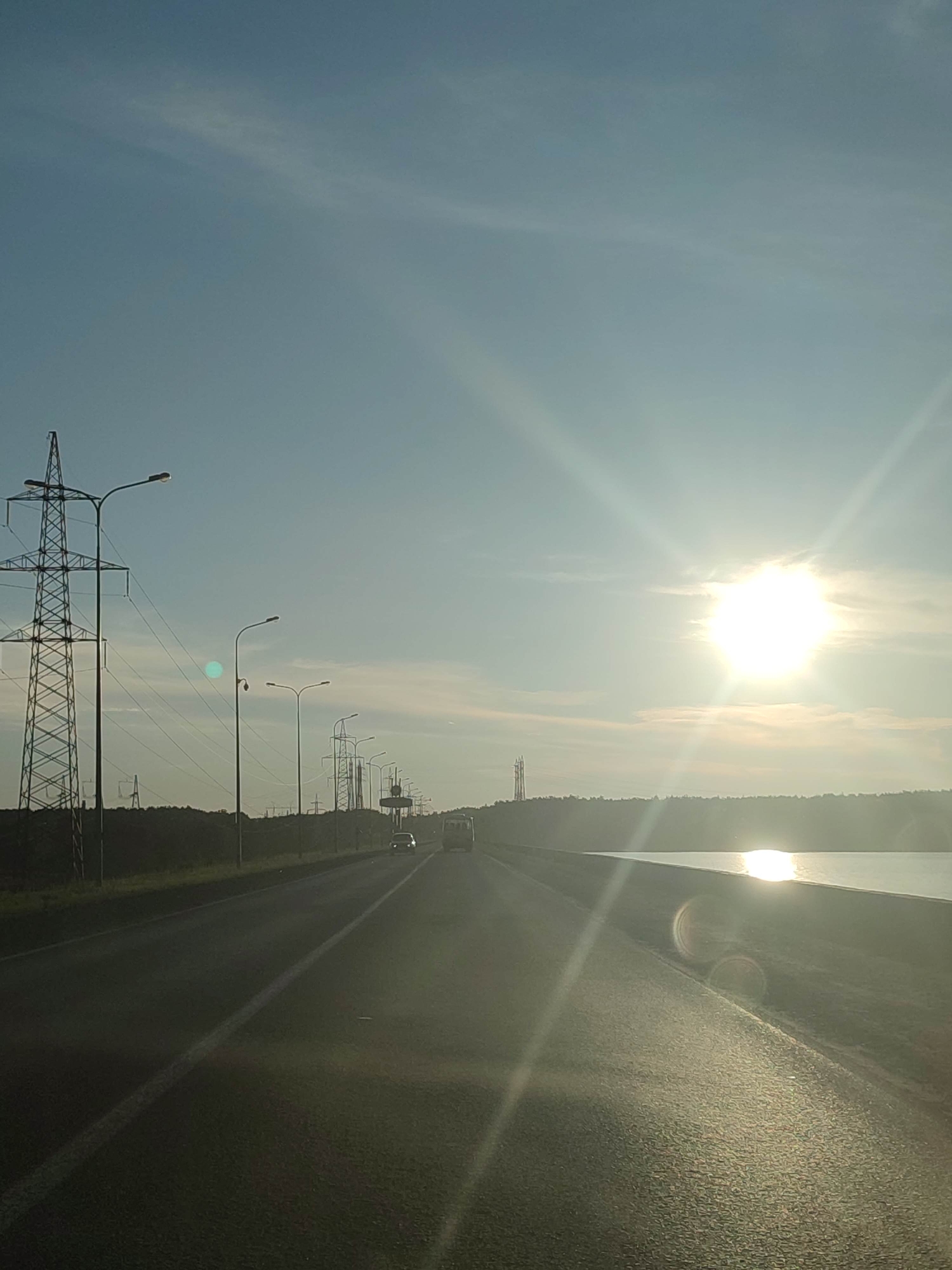This media file captures a scenic view of a road alongside a body of water, likely a lake or reservoir, during what appears to be a sunset or sunrise. The sun is a prominent feature, shining brightly in the upper right quadrant of the image, casting a warm glow and creating lens flares that streak across the frame. The sky is a clear, pale blue with wisps of thin clouds, suggesting fair weather.
The road, paved and marked with a white line, stretches into the distance, curving slightly to the right. On the left side of the road, a series of tall, lattice-like electrical pylons and streetlights are visible, indicating the presence of infrastructure. Trees and other vegetation form a dark line along the horizon behind the pylons. A few vehicles, including a car and a van or bus, are seen driving on the road in the distance.
The body of water to the right of the road reflects the bright sunlight, creating a shimmering effect. The overall mood of the image is serene and tranquil, evoked by the natural beauty of the landscape and the soft light of the sun. There is no visible text in the image. The setting appears to be a rural or semi-rural area, characterized by the open road, power lines, and the expansive natural landscape.