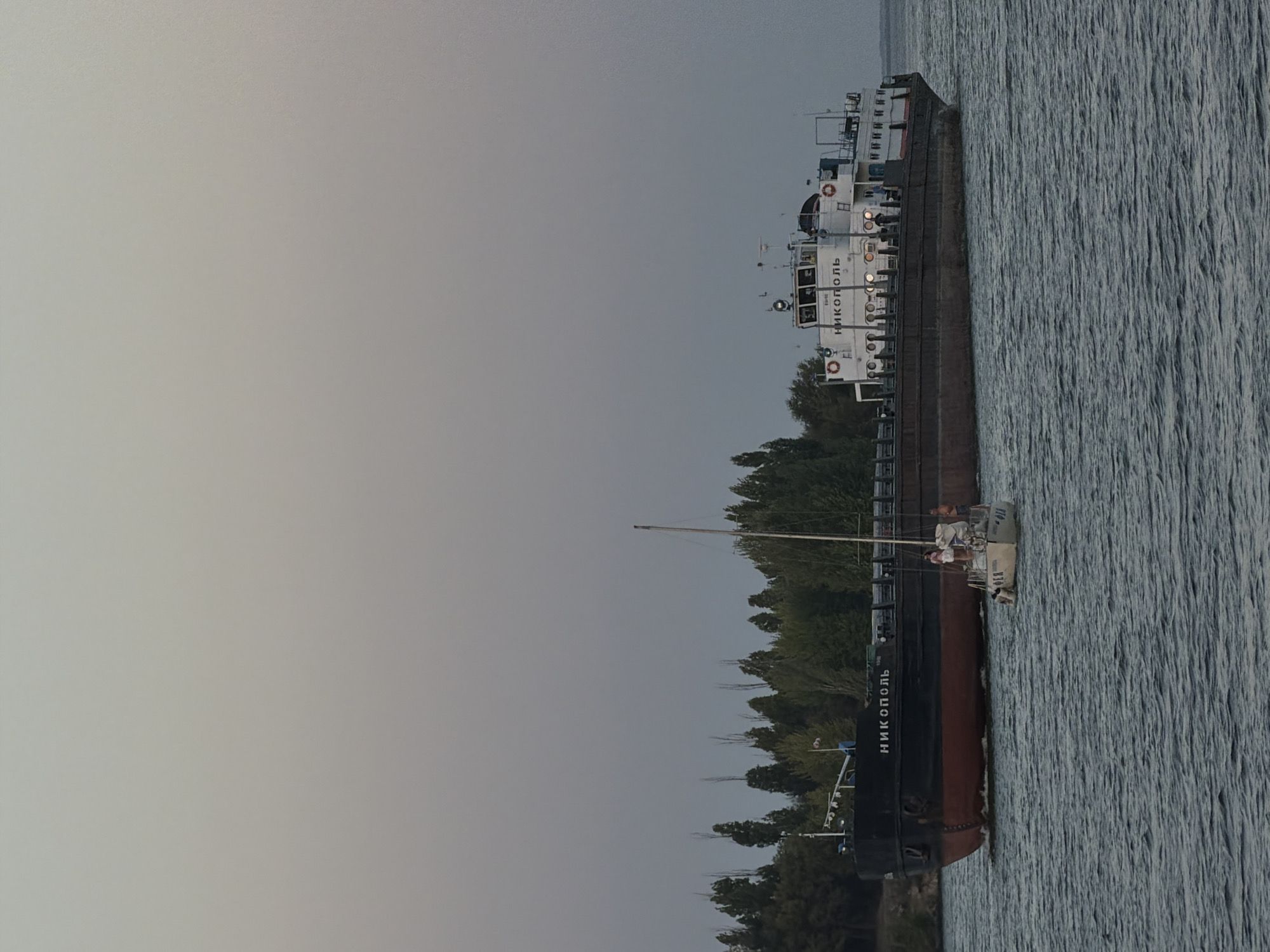 The image shows a wide river with a cargo barge and a sailboat moving on it. The cargo barge, labeled "НИКОПОЛЬ," is dark with a red hull and is the larger of the two vessels. The sailboat is smaller and white, with its sail partially furled. There are a few people visible on the deck of the sailboat and on the barge. The background features a dense line of trees and a pale, overcast sky. The scene appears to be a calm day on the water, possibly during early morning or late evening given the soft light. The location is the Volga River in Kamyshin, Russia, a city known for its port and industrial activity along the river.