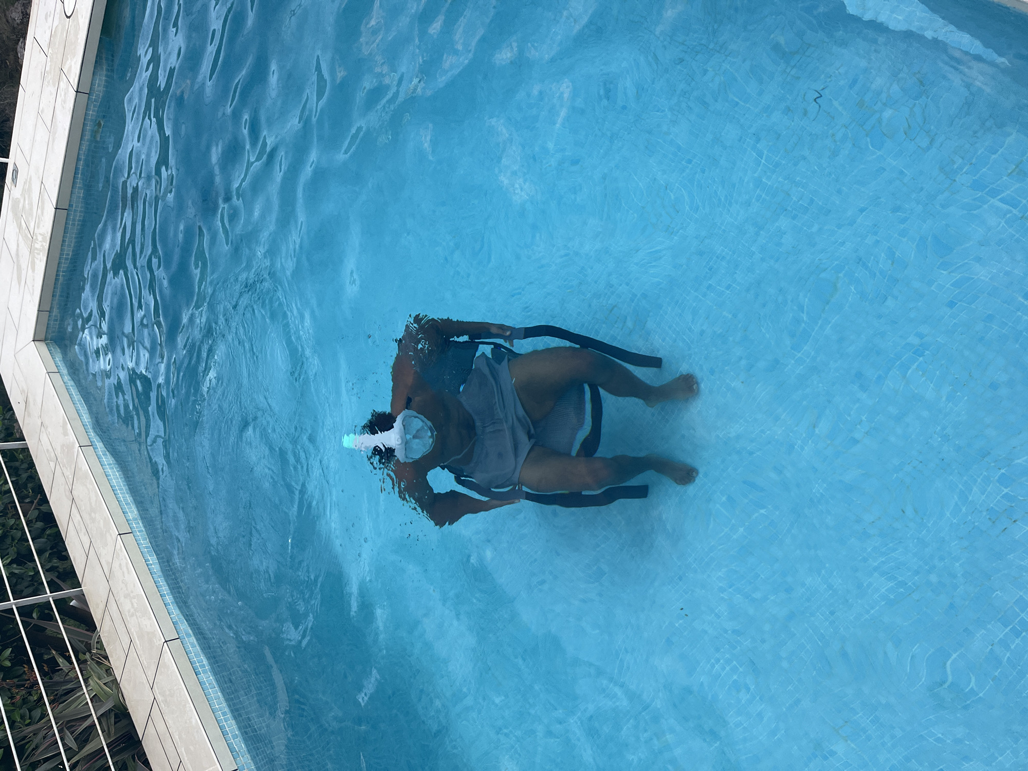 A person wearing a snorkel mask, grey top, and swimming trunks is submerged in a pool of light blue water. They are holding onto a black swimming aid or similar equipment with both hands. The pool has light blue tiled walls. At the edge of the pool on the upper left of the frame, white coping stones are visible next to a small area with green vegetation and white fencing. The location is likely El Poble Nou de Benitatxell / Benitachell, Spain.
