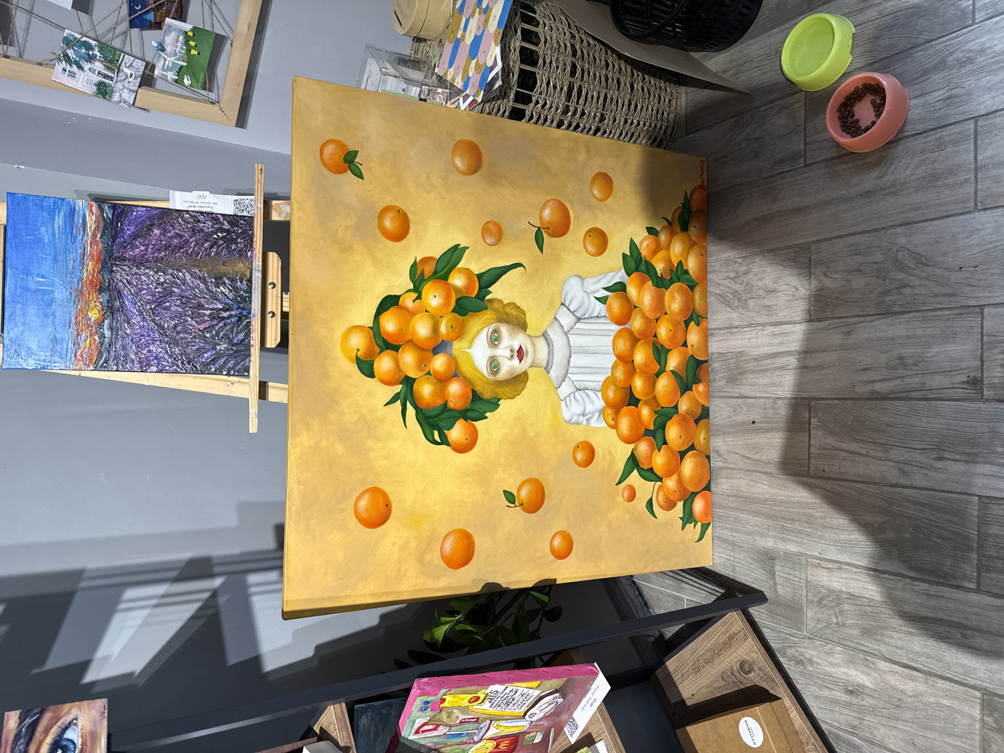 A horizontally-oriented, eye-level photograph captures a room, likely an art gallery or shop, in Batumi, Georgia. The foreground exhibits a diverse array of objects, including paintings, woven baskets, and pet bowls, indicative of an artful or retail space.
Dominating the central composition is a large painting featuring a pale-skinned woman surrounded by oranges on a golden background. This artwork is placed on a shelf or table, alongside other smaller paintings. One of these smaller paintings, positioned to the left on an easel, depicts a landscape with blue and purple hues.
In the top right corner of the image, there are pet bowls in pink and green. Nearby, a woven basket adds a textural element to the scene.
The environment is indicative of a modern setting, with light-colored, wood-like flooring, and gray walls that serve as a backdrop for the artwork. The lighting is bright and even, illuminating the details of the various objects and paintings in the room. The scene is static, devoid of human activity, and appears to be a display or arrangement within the gallery or shop.