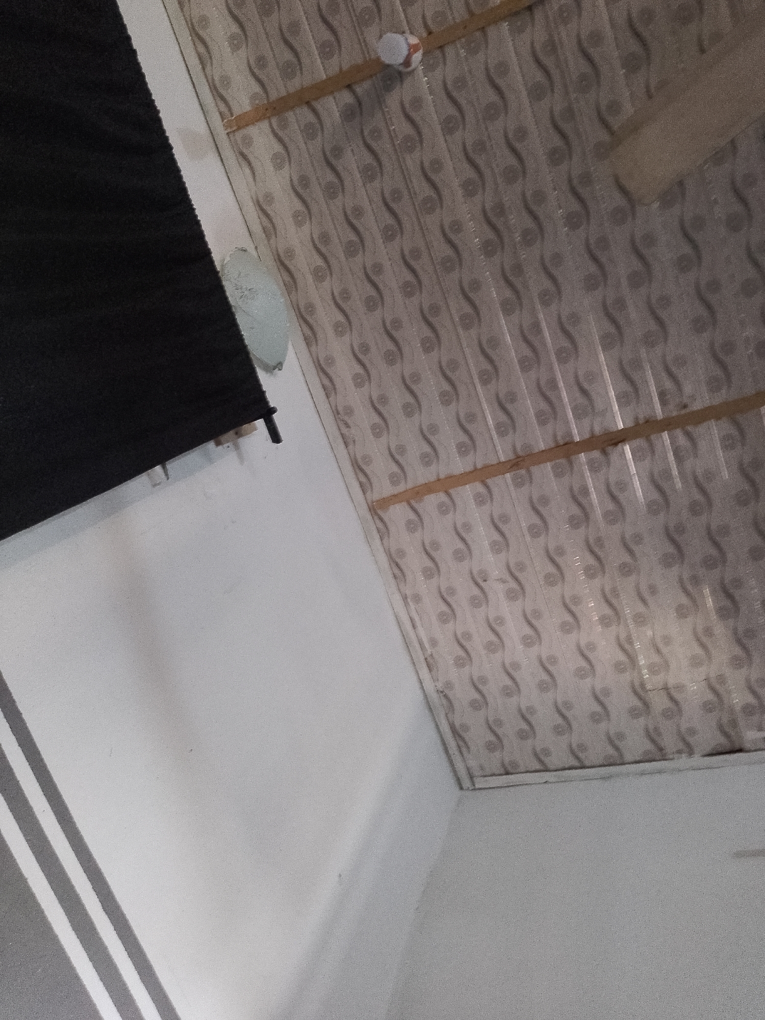 The image depicts an indoor scene with a patterned ceiling. The ceiling has a repeating wavy pattern in light and dark gray. Two wooden planks are visible, horizontally positioned across the ceiling panels. A small, round white object with a red stripe is attached to the ceiling in the upper left quadrant. To the left of the ceiling, a black rolled-up blind or curtain is attached to the wall. The wall itself is painted white and has diagonal gray stripes on the lower left side of the image. The overall perspective suggests the photo was taken from a low angle, looking upwards. The scene is devoid of any people or explicit actions. The location is described as Jalingo, Nigeria.