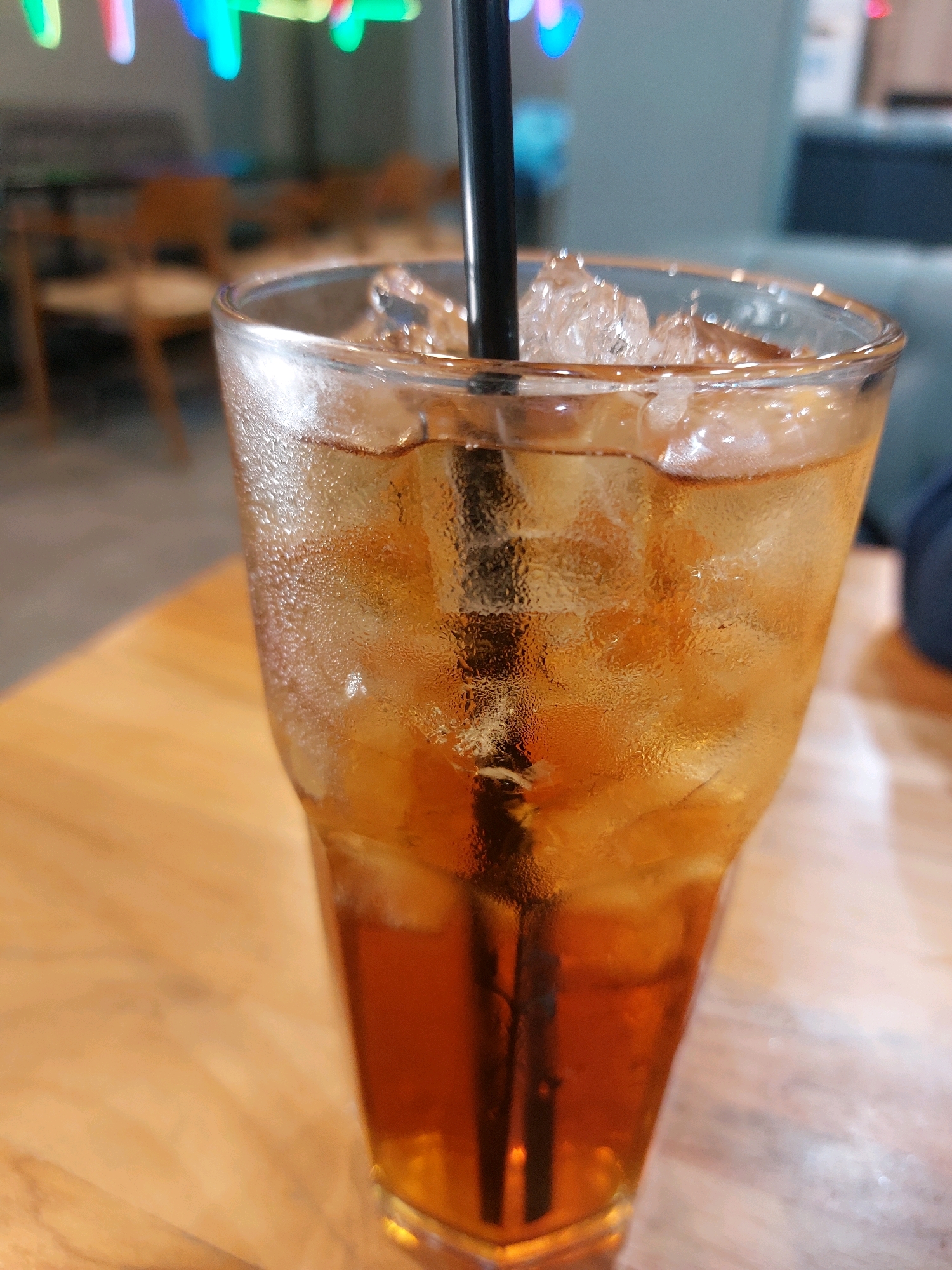 A close-up, eye-level shot focuses on a tall glass filled with iced tea and a black straw. The glass is sweating, indicating the beverage is cold, and sits on a wooden table. In the blurred background, neon lights and out-of-focus furniture suggest an indoor seating area, possibly a cafe or restaurant. The location is identified as Tarakan, Indonesia.