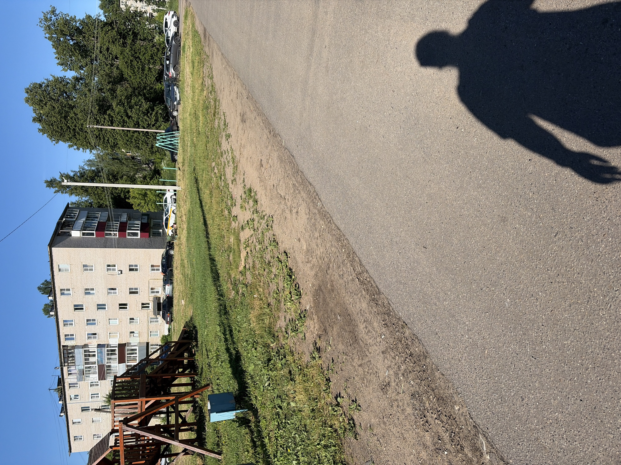 A shadow of a person, facing left, is cast on a paved asphalt path in the foreground of the image. The path runs diagonally from the bottom right to the top left. To the left of the path is a strip of dirt and grass. In the background, a multi-story apartment building with light-colored brickwork and balconies is visible. A children's playground structure, made of wood, is located to the left of the building. Several cars are parked in front of the building and along the side of the path. Tall green trees are situated behind the apartment building and playground. The sky is clear and blue. The scene appears to be a residential area in a city, likely on a sunny day.