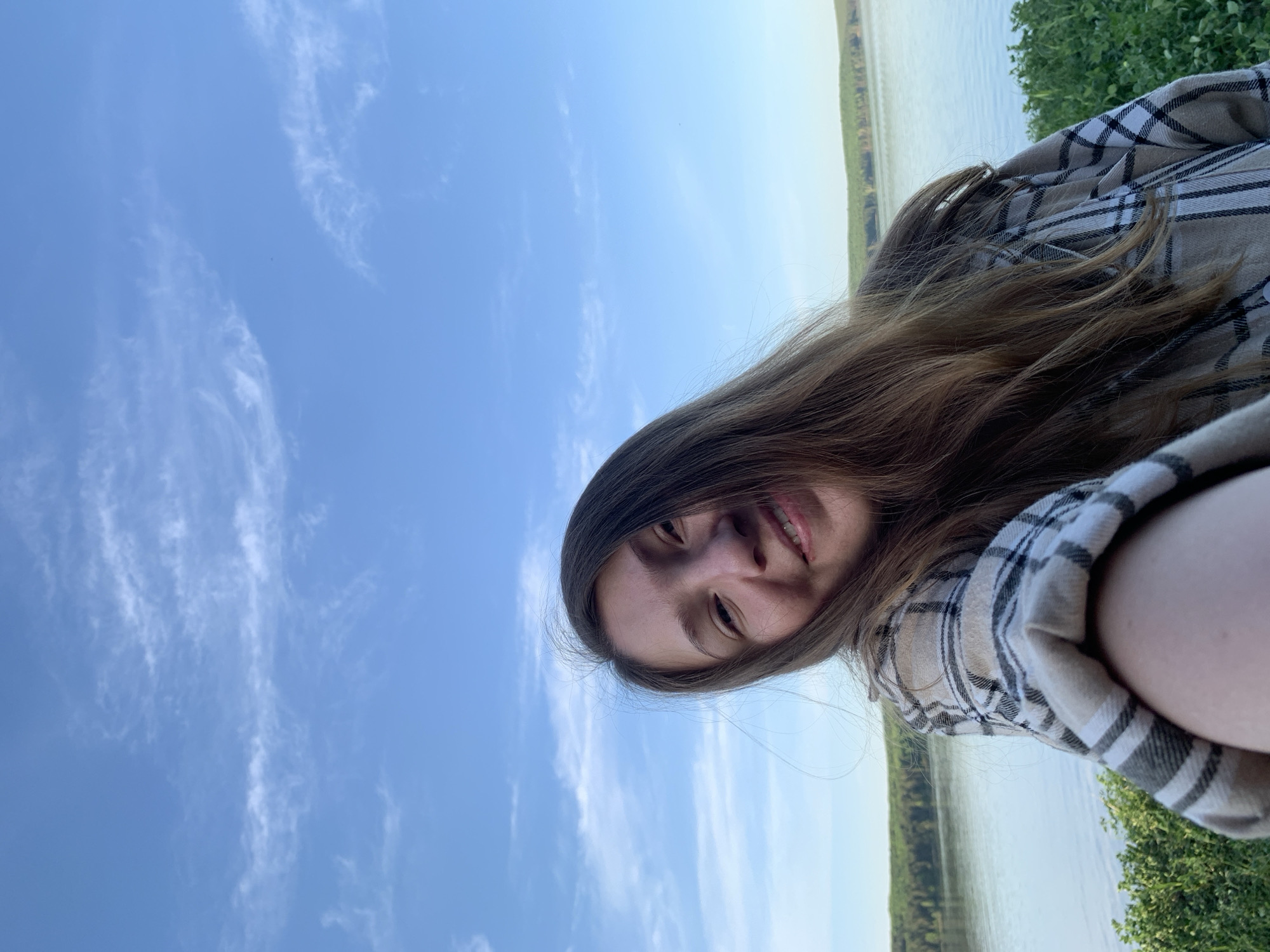 A selfie image shows a woman with long brown hair and a plaid shirt. The background consists of a blue sky with wispy clouds, a body of water, and some green foliage. The location is Chaikovsky, Russia.