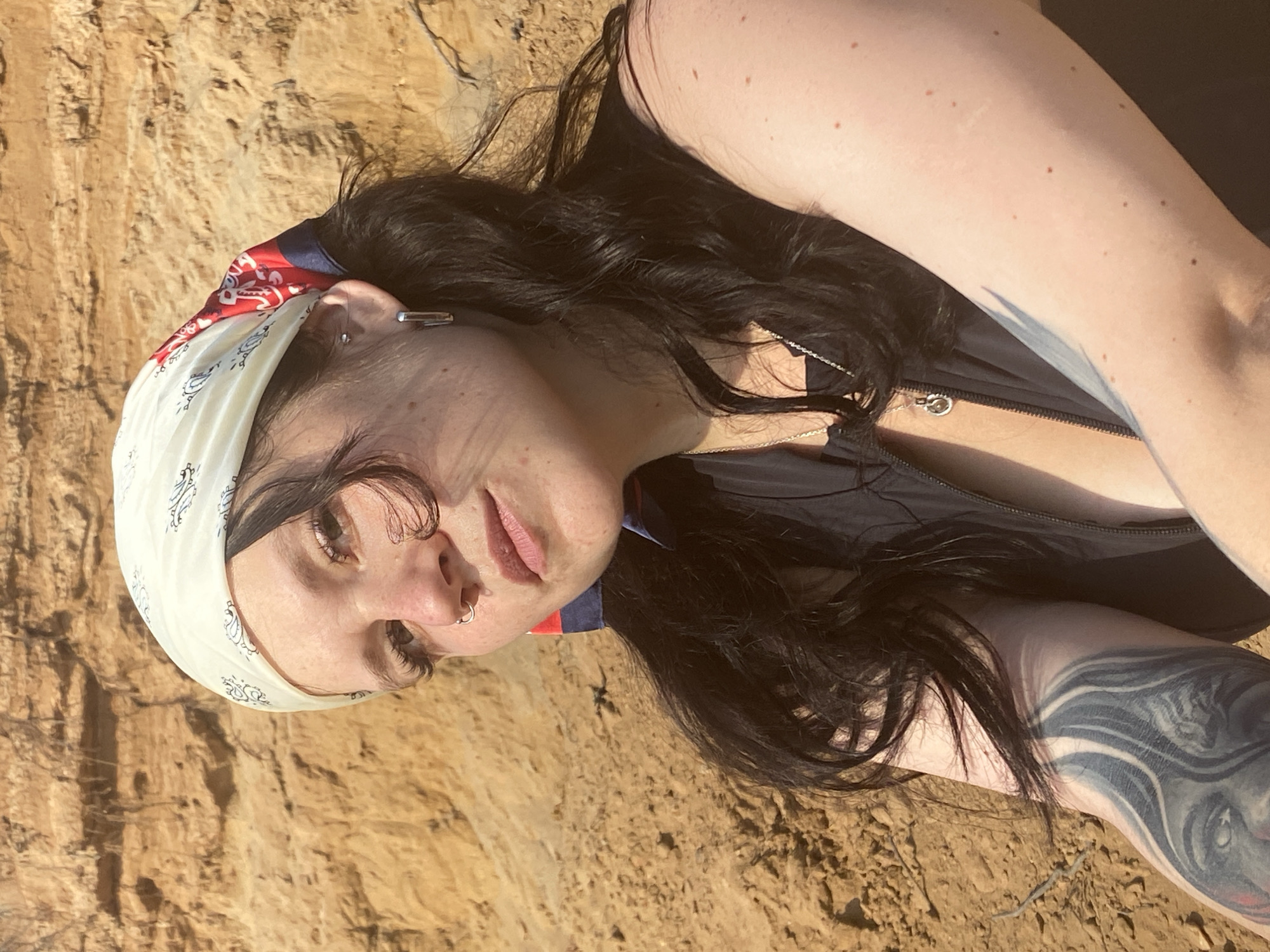 In Novyi, Russia, a young woman with dark hair is captured in a close-up portrait against a backdrop of sandy cliffs. She wears a cream-colored bandana with a black pattern and red and blue accents, a dark sleeveless top, and a silver necklace with a pendant. Her left arm features a large, detailed tattoo. The lighting suggests an outdoor setting, likely during the day, with the sun casting a warm glow.