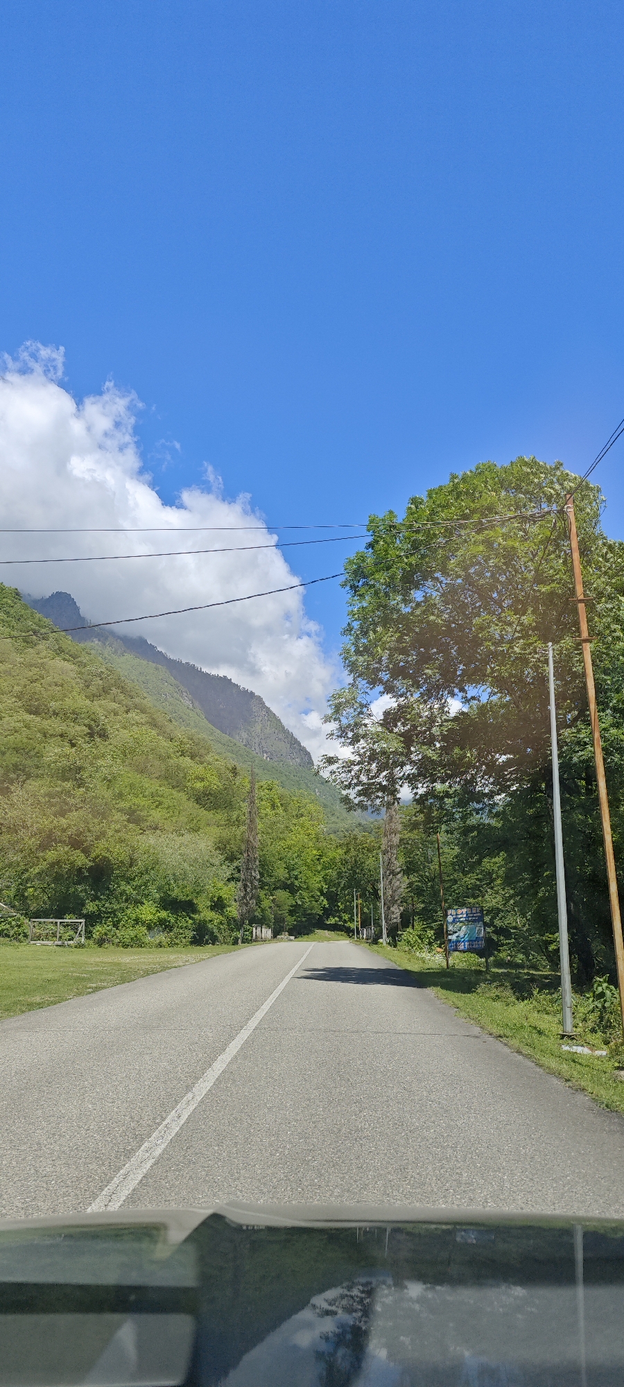 The image presents a first-person view from inside a vehicle, looking forward along a paved road winding through a scenic mountainous and forested landscape. The asphalt road features a clear white solid line down its center.
On both sides of the road, dense green trees and vegetation line the path. In the distance and background, steep, verdant mountains rise, their upper slopes partially obscured by large white clouds against a vibrant blue sky. Utility poles and power lines are visible, with a prominent tall wooden pole situated on the right side of the road. Another thinner, lighter-colored pole stands further along on the right. A blue sign with white text and graphics is partially visible on the right, nestled among the trees.
The scene is captured on a bright, sunny day, likely late morning or early afternoon, with scattered clouds. Reflections of the sky, clouds, and surrounding foliage are visible on the vehicle's dashboard and windshield at the bottom of the frame. The overall impression is one of a journey through a serene natural environment, typical of mountainous regions like Bzypta, Abkhazia. The text on the blue sign is not clearly legible.