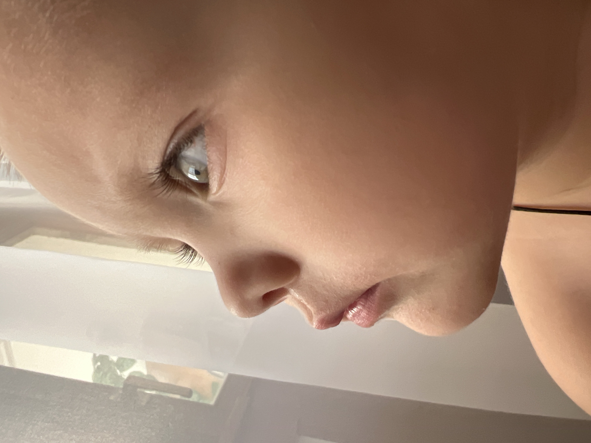 A close-up, tilted shot shows the side profile of a child's face. The child has fair skin, light-colored eyelashes, and a visible eye reflecting light. The nose is small and slightly upturned, and the lips are pink with a hint of gloss. Part of the child's neck and shoulder are visible. The background is blurred, but shows a window with a view of greenery outside. There is no information available to determine if this picture was taken in Марковский, Russia.