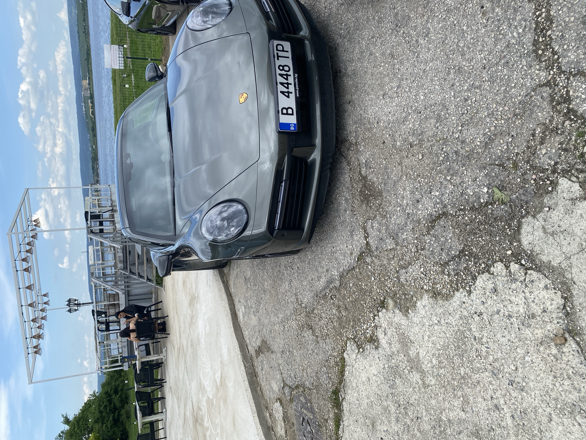 The image shows a gray Porsche parked next to a cafe. The license plate number is B 4448 TP. Two women are seated at a table in the cafe. The background includes a body of water and the sky with clouds. The scene is likely located in Konstantinovo, Bulgaria.