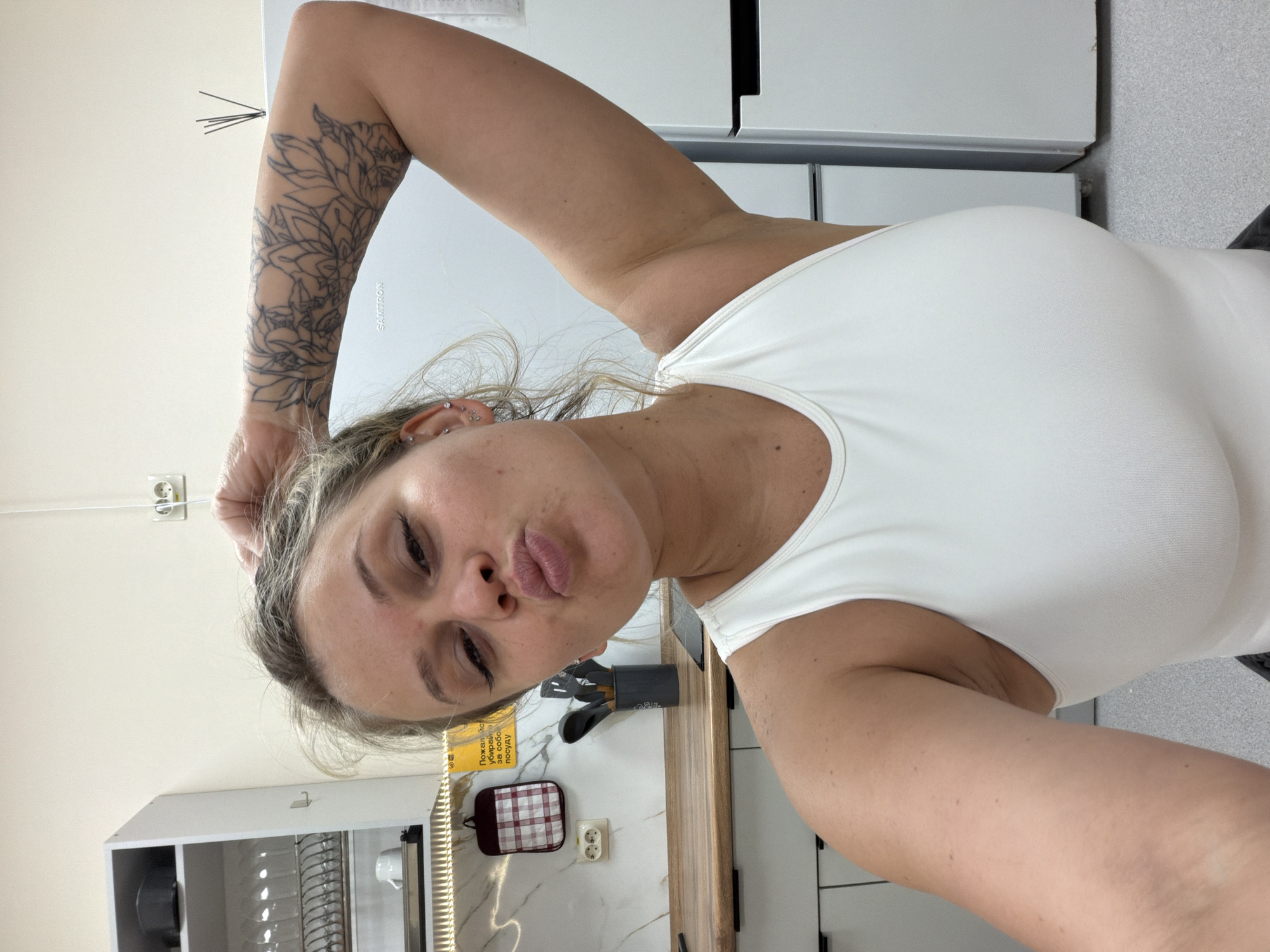 A woman is posing for a selfie in a kitchen. The kitchen has white cabinets and a marbled countertop. Kitchen utensils are visible on the countertop. The woman is wearing a white top and has a tattoo on her left arm. She is making a kissing face at the camera. There is a yellow sign on the wall. The location is Sarapul, Russia.