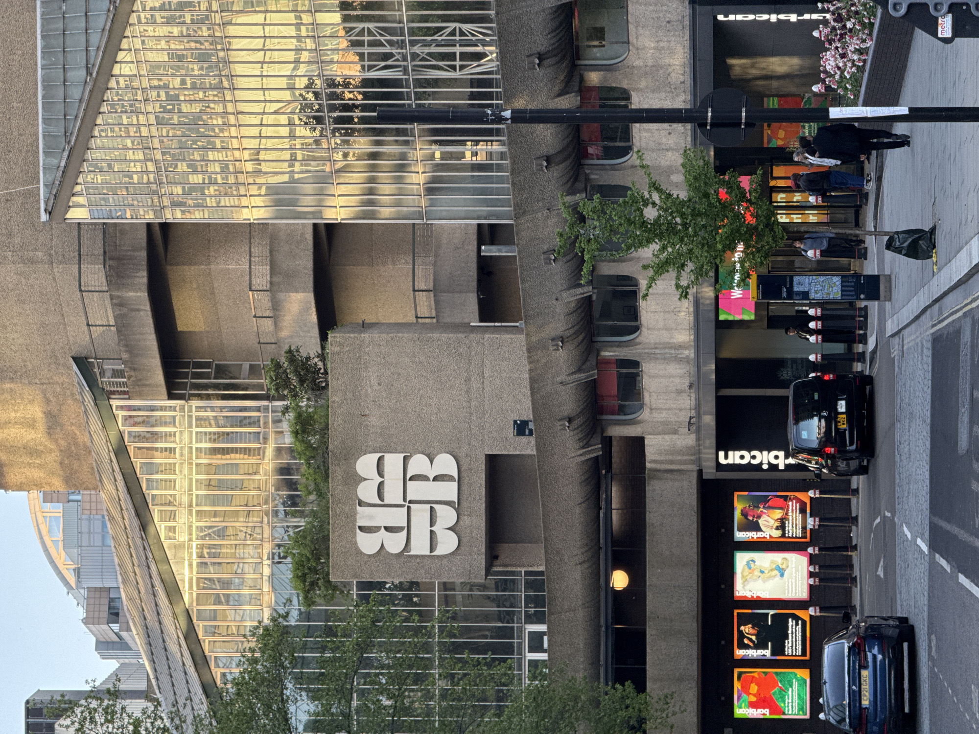 The image shows the Barbican Centre in London, United Kingdom. The architecture is predominantly Brutalist, featuring exposed concrete and geometric shapes. The Barbican logo is visible on a concrete block. A modern glass-faced building reflects the golden light of the setting sun. A street runs along the building's edge, with a bus shelter, street signs, posters advertising Barbican events, parked cars, and pedestrians.