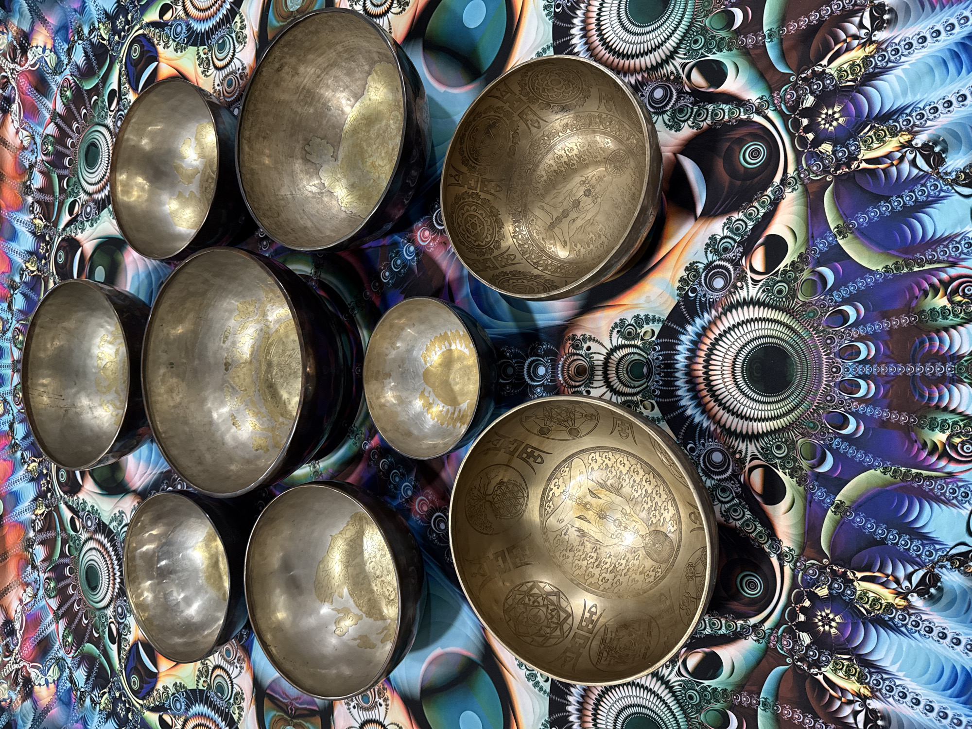 The image shows nine singing bowls of varying sizes arranged in a roughly rectangular grid on a kaleidoscopic patterned surface. The bowls are metallic, ranging in color from silver to gold. Some of the bowls have intricate designs etched into their interiors. The background pattern features fractal designs with repeating circular and teardrop shapes in shades of blue, purple, green, and yellow. No people are visible in the image. The location is unknown.