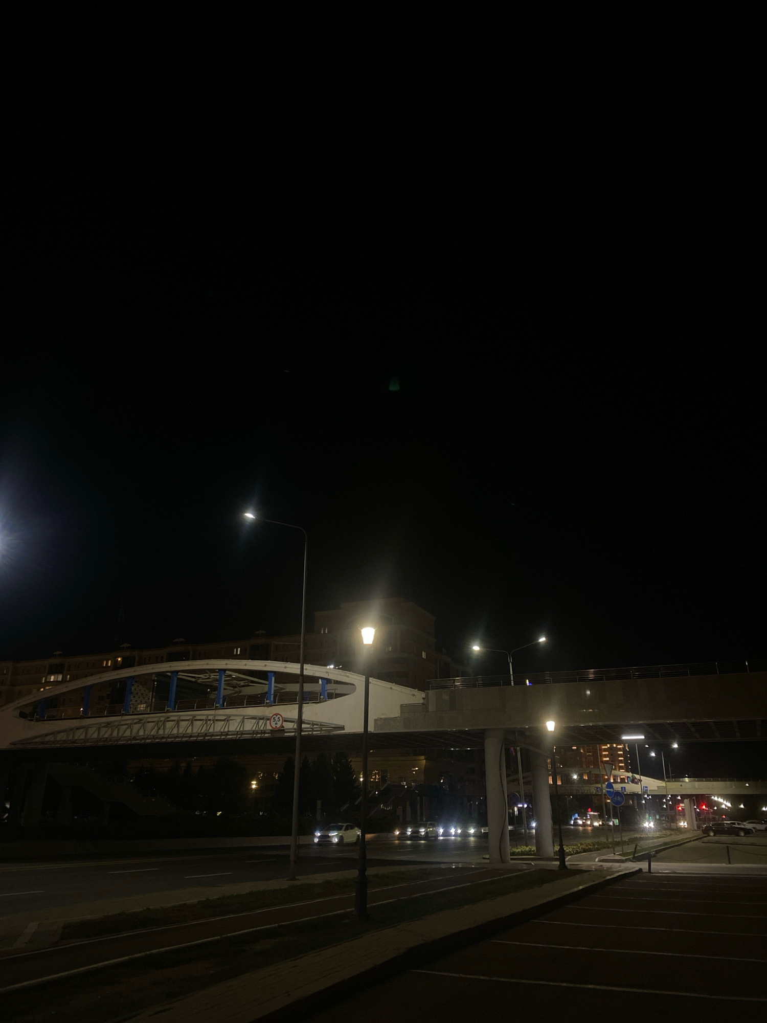 The image is a nighttime scene in Astana, Kazakhstan, featuring an elevated pedestrian bridge illuminated by streetlights. The bridge, with its distinctive curved white railings and blue structural supports, spans across a road where vehicles with their headlights on are visible. Buildings with illuminated windows can be seen in the background, suggesting an urban environment. Streetlights are strategically placed along the road and near the bridge, casting pools of light. The foreground shows a paved area, possibly a parking lot or sidewalk, with marked lines. The sky is dark, indicating it is late at night.