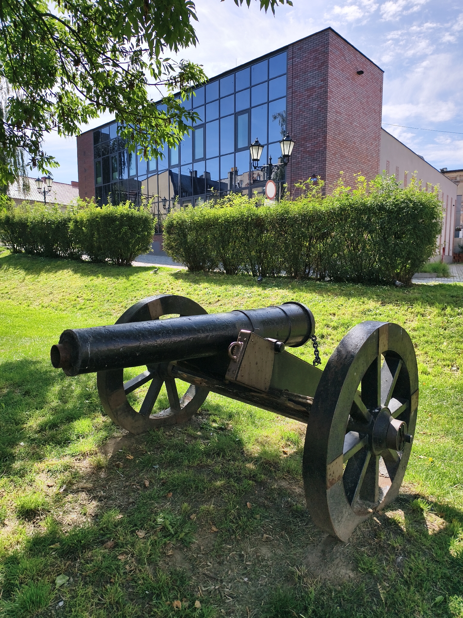 An antique black cannon on a wooden carriage with two large spoked wheels is positioned on a grassy lawn. The cannon is facing left in the frame, and its barrel is visible from the breech to the muzzle. Behind the cannon, a manicured hedge forms a boundary. Further in the background, a modern brick and glass building stands, with its glass facade reflecting the sky and surrounding environment. Two ornate lampposts are visible in front of the building, along with a circular sign. Trees with green foliage overhang the scene from the left. The ground is covered in green grass, with patches of dirt and fallen leaves. The sky is blue with scattered white clouds. The location is the area in front of the Tczew Museum in Tczew, Poland.