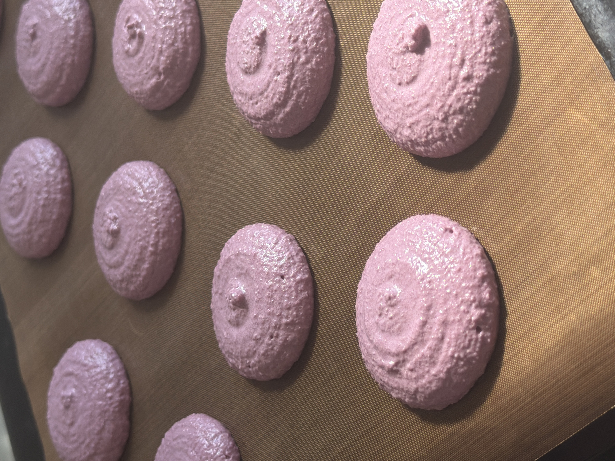 The image shows a close-up of several pink macarons arranged on a brown baking mat. The macarons are circular and appear slightly textured. The scene is indoors. There is no indication that this image is related to Tel-Aviv, Israel. There are no people visible.