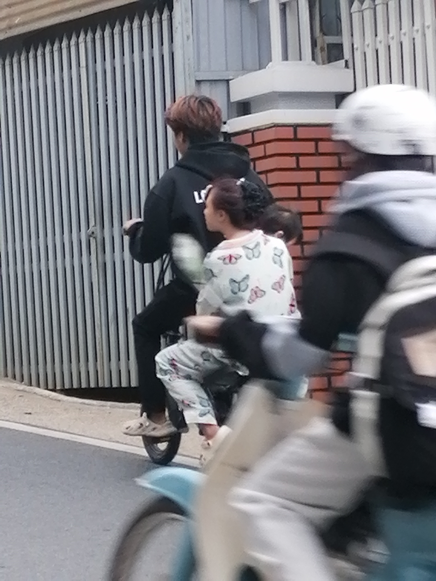 The image shows three people on two motorbikes, moving along a paved road in Da Lat, Vietnam. The primary focus is on a motorbike with two people. The person in the back, wearing a black hoodie and black pants, is driving. They have reddish-brown hair. The person in the front, sitting behind the driver, wears a white long-sleeved shirt with a pink and red butterfly pattern and light-colored pants. They have dark hair pulled back. Their feet are visible, wearing light-colored slip-on shoes. A third person, mostly out of focus due to motion blur, is on another motorbike in the foreground, wearing a white helmet and a grey jacket.
The background features a grey metal gate with vertical bars, and a brick wall with orange bricks. The scene appears to be outdoors, on a street, likely during the day, given the lighting. The weather is unclear, but the general atmosphere suggests a typical urban or suburban setting. The activity is simply transportation by motorbike. There is no visible text in the image.