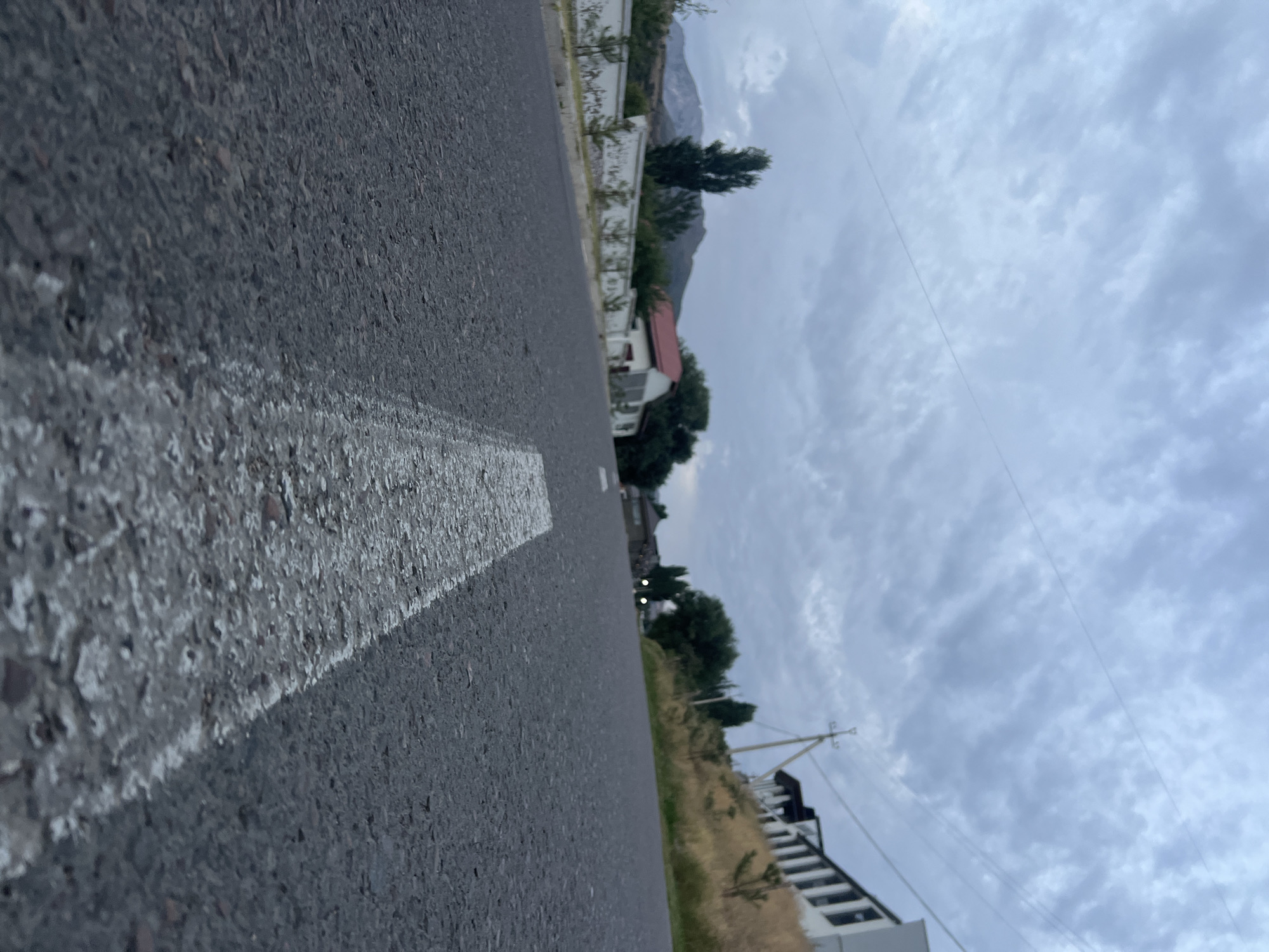 The image depicts a low-angle view of an asphalt road with a white lane marking, looking upwards towards a cloudy sky. In the background, there are buildings, trees, a white wall, and mountains. The scene appears to be in Yusuphana, Uzbekistan, given the architectural style and natural landscape. There are no people or animals visible in the image. The overall atmosphere is overcast and subdued, suggesting it might be evening or a cloudy day.
