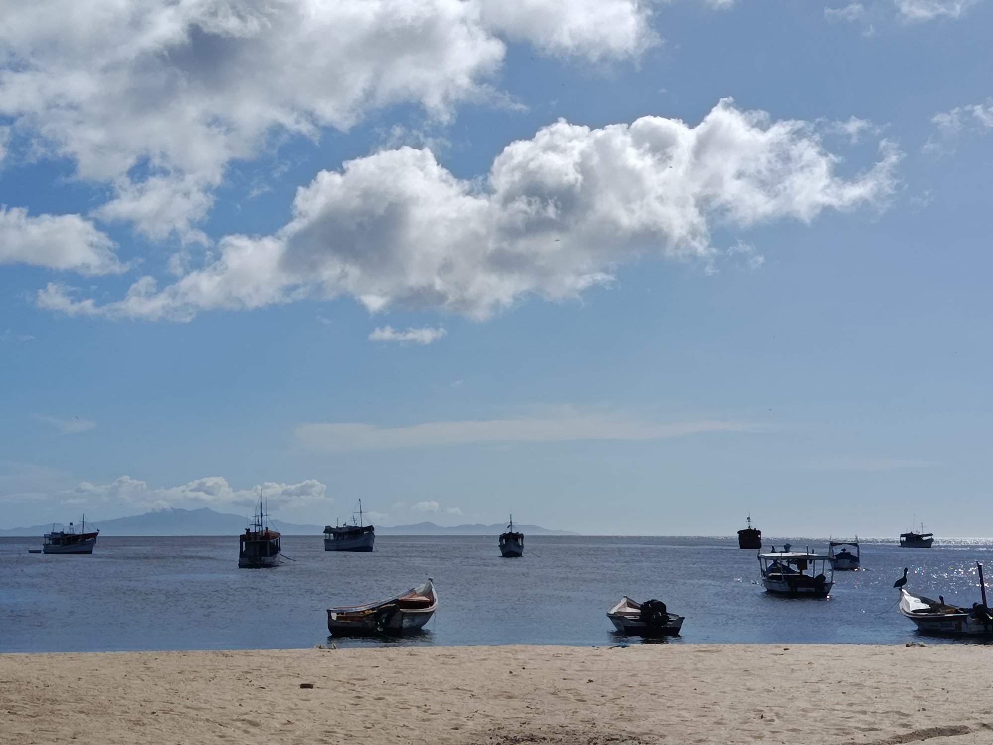 The image depicts a tranquil scene at the beach in Juan Griego, Venezuela. The foreground features a sandy beach leading to calm, blue water. Several small to medium-sized boats are scattered across the water, some moored closer to the shore and others further out. In the background, a distant shoreline with mountains under a bright, partly cloudy sky is visible. The overall impression is one of a peaceful coastal setting.