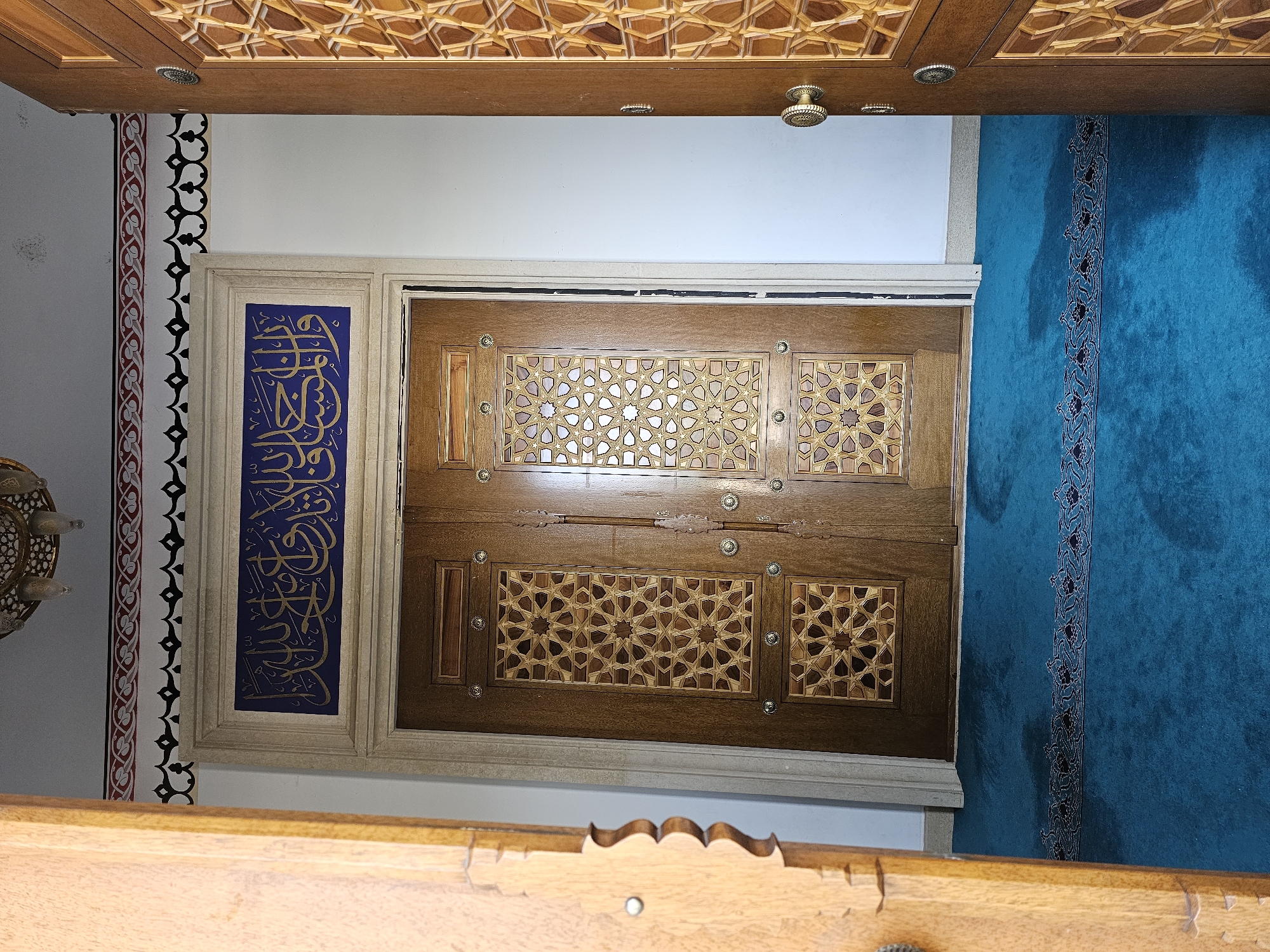 This image depicts an interior view of what appears to be a mosque in Özgön, Kyrgyzstan. The focal point is a set of ornate wooden double doors, featuring intricate geometric carvings and decorative metal accents. Above the doors, a rectangular blue plaque displays Arabic calligraphy in gold lettering. The doors are flanked by a plain white wall and a partially open wooden door on the right, also decorated with geometric patterns. The floor is covered with a vibrant teal carpet, adorned with a decorative border pattern. Above the doors, a frieze with red and black patterned trim runs along the wall, leading up to a light fixture or chandelier. The overall impression is one of traditional Islamic architectural and decorative style.