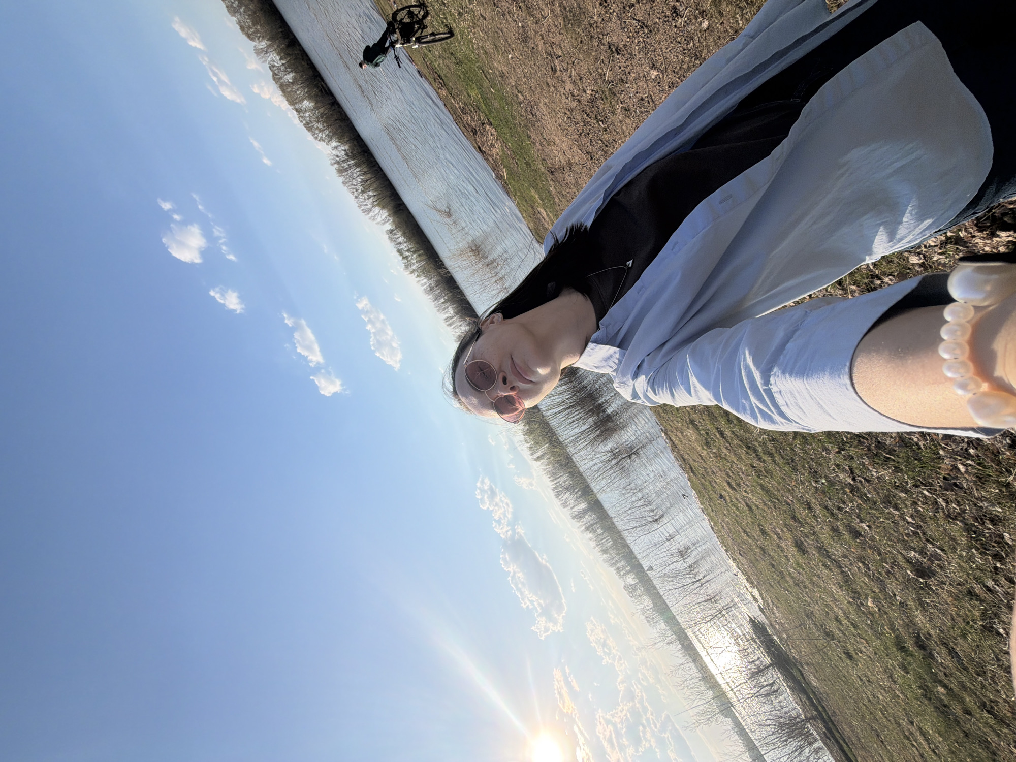 A person is holding the camera at arm's length, angled upward to capture a selfie in Chaikovsky, Russia. The person is wearing a light-colored button-down shirt over a dark top, and round sunglasses. A pearl bracelet is visible on their wrist. Behind the person, the backdrop consists of a field with grass, a body of water reflecting sunlight, and a line of trees along the water's edge. In the upper left corner, there's a wooden barrier or boardwalk with a bicycle resting on it. The sky is blue with some clouds, and the sun is shining brightly, causing glare.