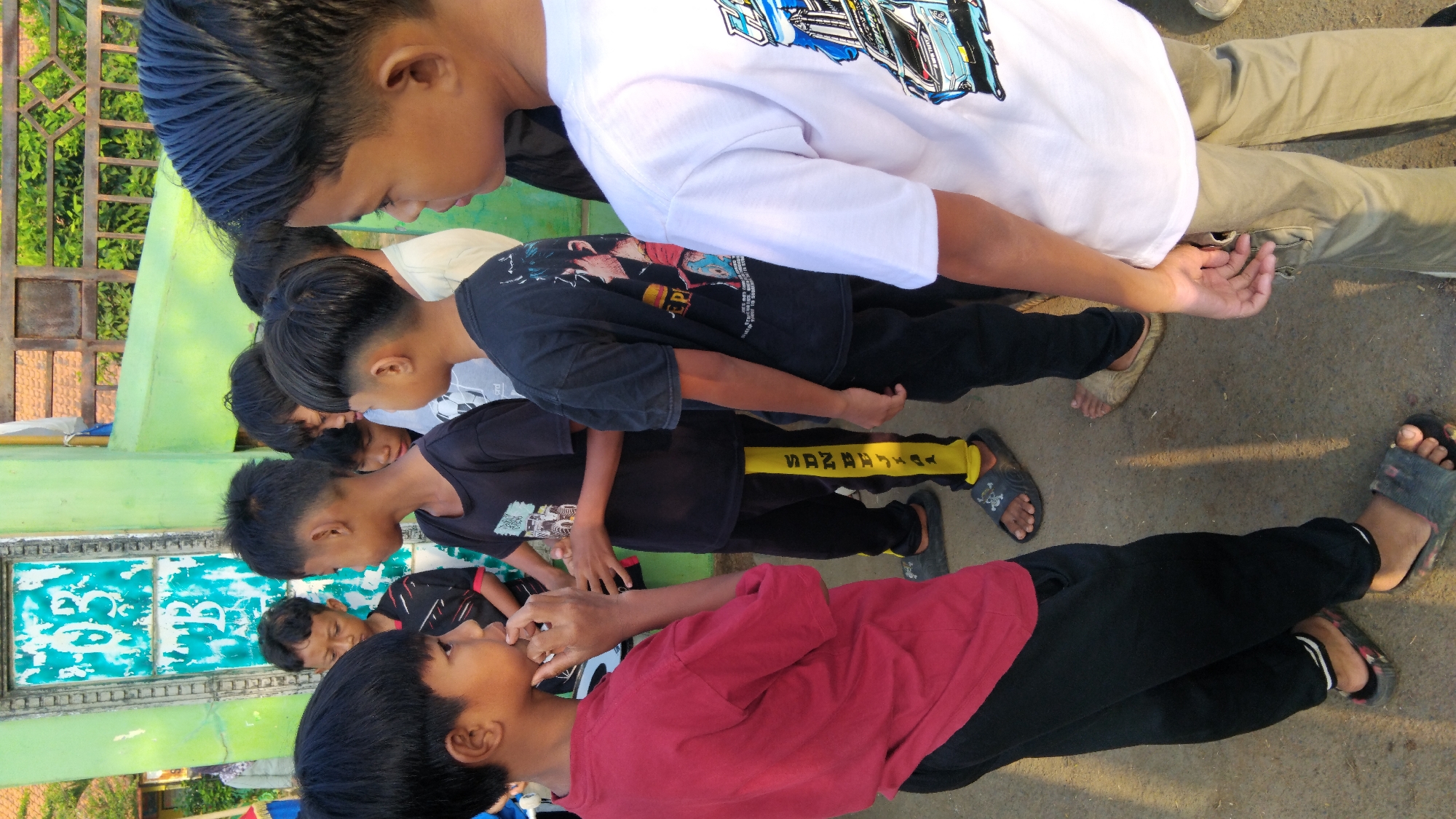 In Sembojo, Indonesia, a group of young boys stand in front of a weathered green building with a white-painted door. The boys, dressed in casual t-shirts and pants, appear to be in conversation. The foreground is dominated by the side of a boy wearing a red t-shirt and black pants. To his left, another boy in a black t-shirt and black pants with yellow stripes is visible. Further back, other boys are partially obscured, one in a black t-shirt with an anime-style graphic and another in a white t-shirt. The ground is paved, and a metal fence with wooden planks is visible in the background, suggesting an outdoor, possibly school or community area. Sunlight casts shadows on the ground, indicating it is daytime.