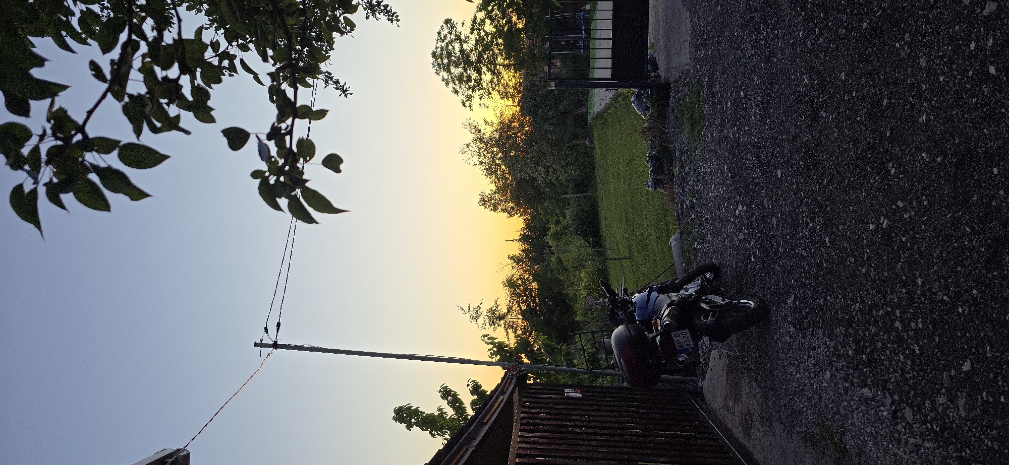 This media file depicts an outdoor scene with a clear sky transitioning to a warm sunset. The image is oriented vertically, with the sky occupying the upper left portion and the ground and objects filling the rest.
**Subjects:**
The main subjects include a motorcycle parked on a gravel surface, a wooden structure resembling a shed or fence, a grassy area, and trees. In the foreground, dark green leaves from a tree branch are visible, silhouetted against the sky. A utility pole with wires also stands out against the sky.
**Activity or Event:**
No specific activity or event is clearly depicted, as the scene appears to be a still moment. The motorcycle is stationary.
**Notable Details:**
The time of day is clearly sunset, indicated by the gradient of the sky from a pale blue to a vibrant orange and yellow. The weather appears to be clear. The presence of the motorcycle and the gravel surface suggest a residential or rural setting, possibly a driveway or yard. The wooden structure adds to this impression. The lighting is characteristic of the "golden hour" before sunset.
**Visible Text:**
On the license plate of the motorcycle, there is visible text that appears to be "BP B 4320". There is also a sticker on the side of the wooden structure.