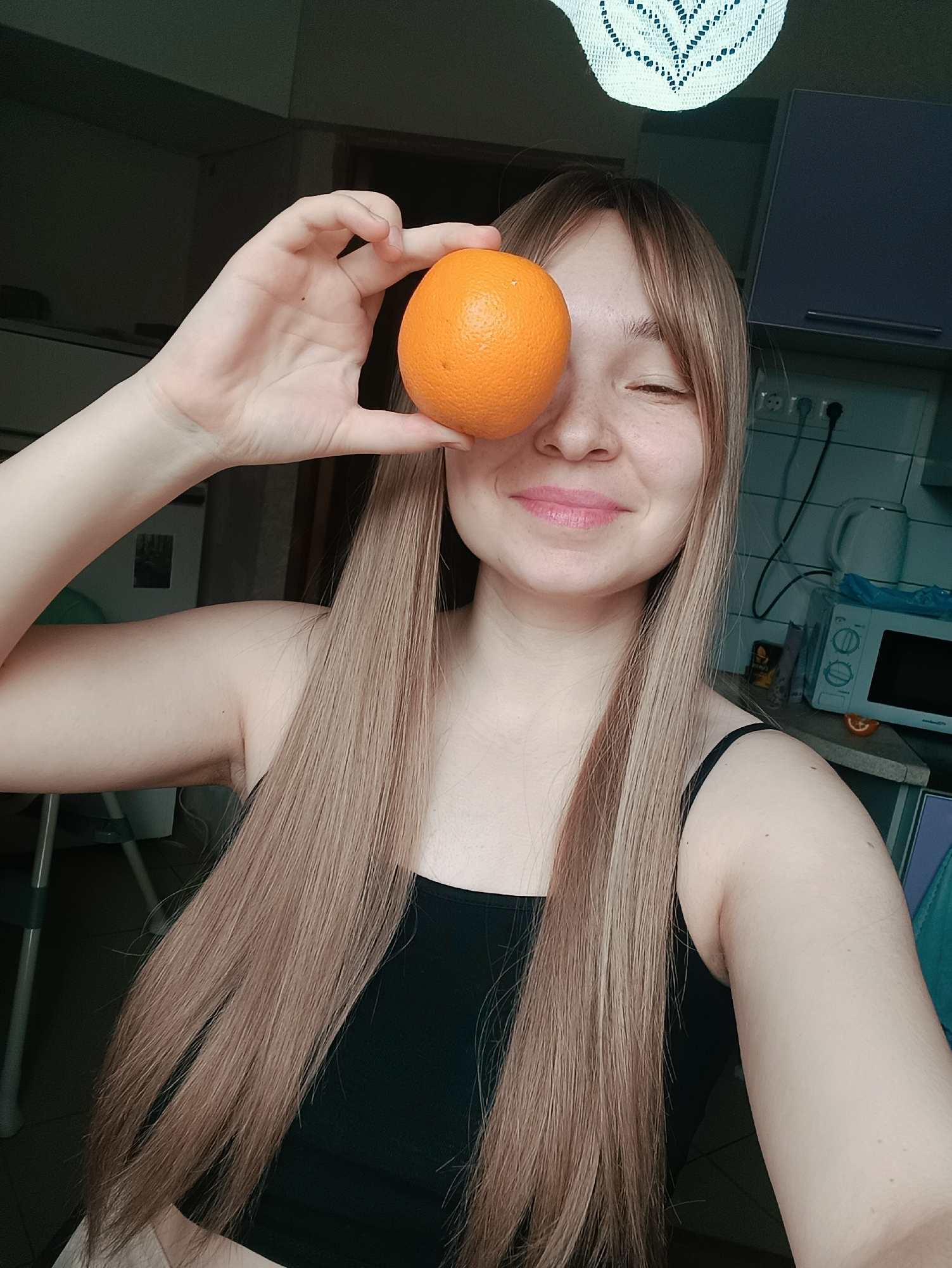 A young woman is holding an orange over her right eye. She is in what appears to be a kitchen. A microwave and kettle are visible in the background. The woman has light-colored hair, fair skin, and freckles. The city of Izhevsk, Russia, cannot be verified based on the visual information provided.