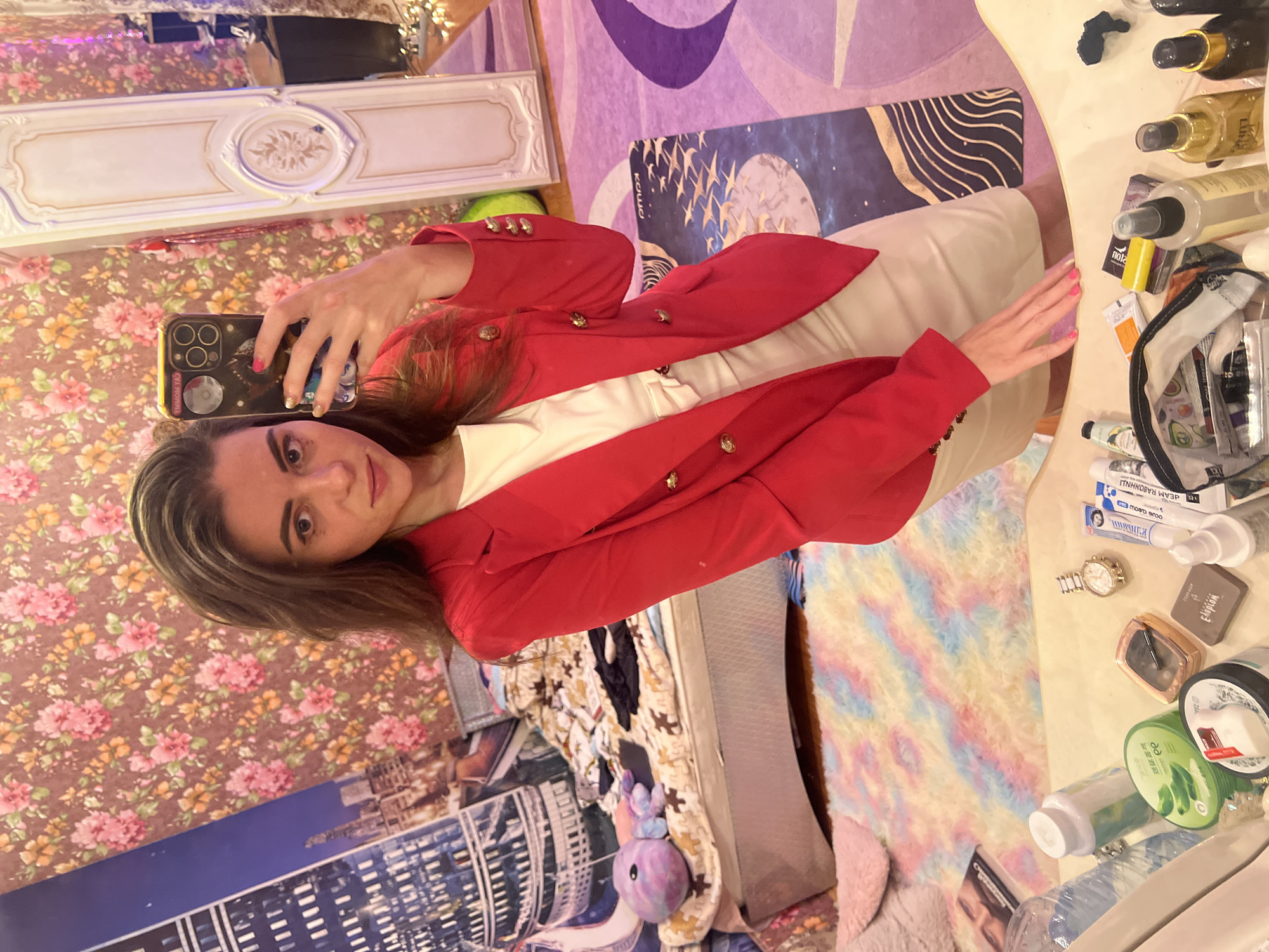 A young woman is taking a mirror selfie in a bedroom. She is wearing a red blazer over a light-colored dress and is holding a smartphone in her right hand. Her left hand rests on a vanity in the foreground, which is cluttered with various cosmetic items and bottles. The background of the room features floral wallpaper and a bed that appears to be unmade. The overall impression is that of a personal space, possibly a bedroom, with decorative elements like a large mirror and a colorful rug. The presence of a smartphone and makeup suggests the woman is in the process of preparing or has just finished doing so. There is no indication of the city of Krasnoe-na-Volge, Russia, in the image.