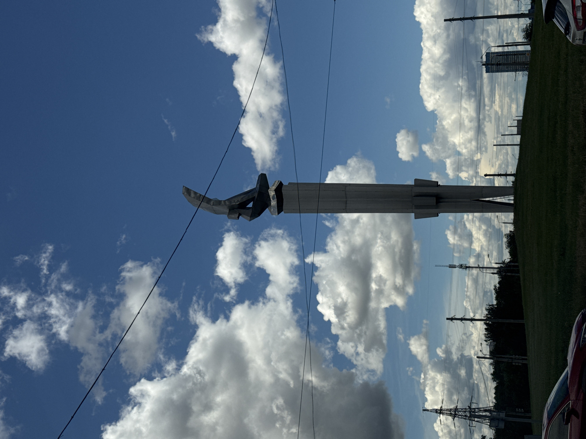 The image shows a monument under a cloudy sky, with power lines crossing the view. The monument is a large, light-colored structure with a hook-shaped element at the top. A grassy area is visible behind the monument, leading to trees and a building in the distance. Parts of cars are visible on either side of the image. The location is the city of Unknown, Unknown.