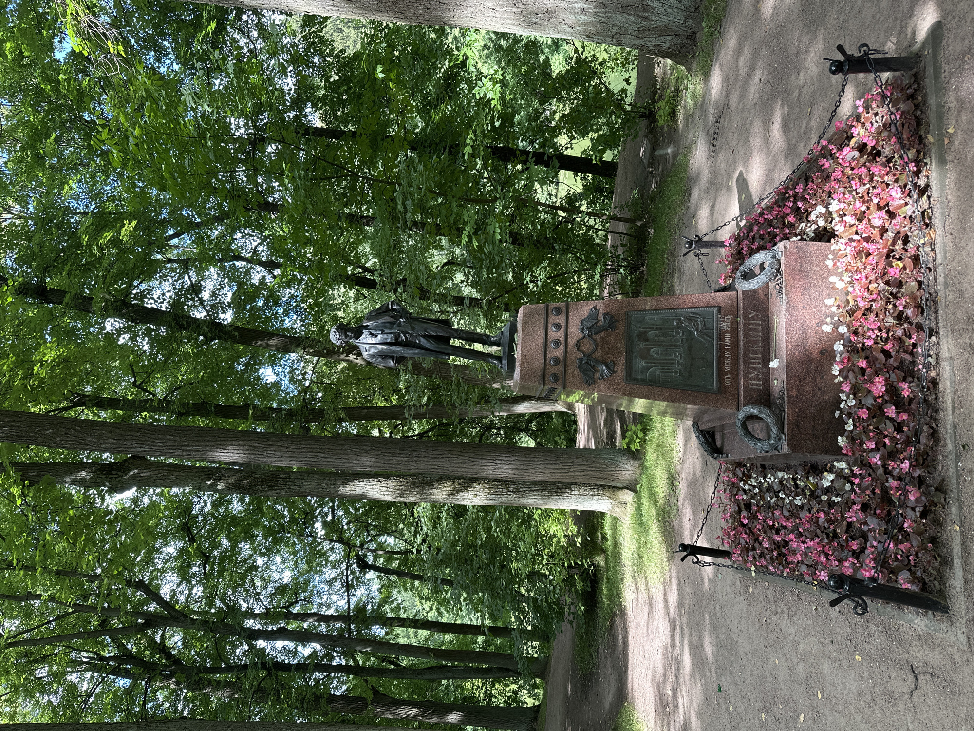 The image captures a monument in a park-like setting, likely a cemetery or historical estate, given the location context of Ostafievo, Russia. The monument itself is a substantial granite structure adorned with a bronze statue of a man in historical attire, possibly a poet or prominent figure, standing in a contemplative pose. The base of the monument features intricate bronze reliefs and inscriptions, though the text is partially obscured. Surrounding the monument is a bed of vibrant pink and white flowers, adding a touch of color and life to the scene. A decorative metal chain fence encloses the flower bed and the monument, with prominent black posts and looped chains.
The setting is a lush, green forest with tall trees casting dappled shadows on the pathway. The sunlight suggests it is daytime, likely late morning or early afternoon, and the weather appears to be pleasant, with clear skies visible through the canopy. The overall atmosphere is serene and solemn, befitting a memorial site. The image's orientation is rotated, with the monument and pathway running vertically. A notable detail is the wreath placed on the monument, a common symbol of remembrance. The visible text on the monument appears to be in Cyrillic script, with a portion reading "ПУШКИНУ", which translates to "To Pushkin". This strongly suggests the monument is dedicated to the renowned Russian poet Alexander Pushkin.