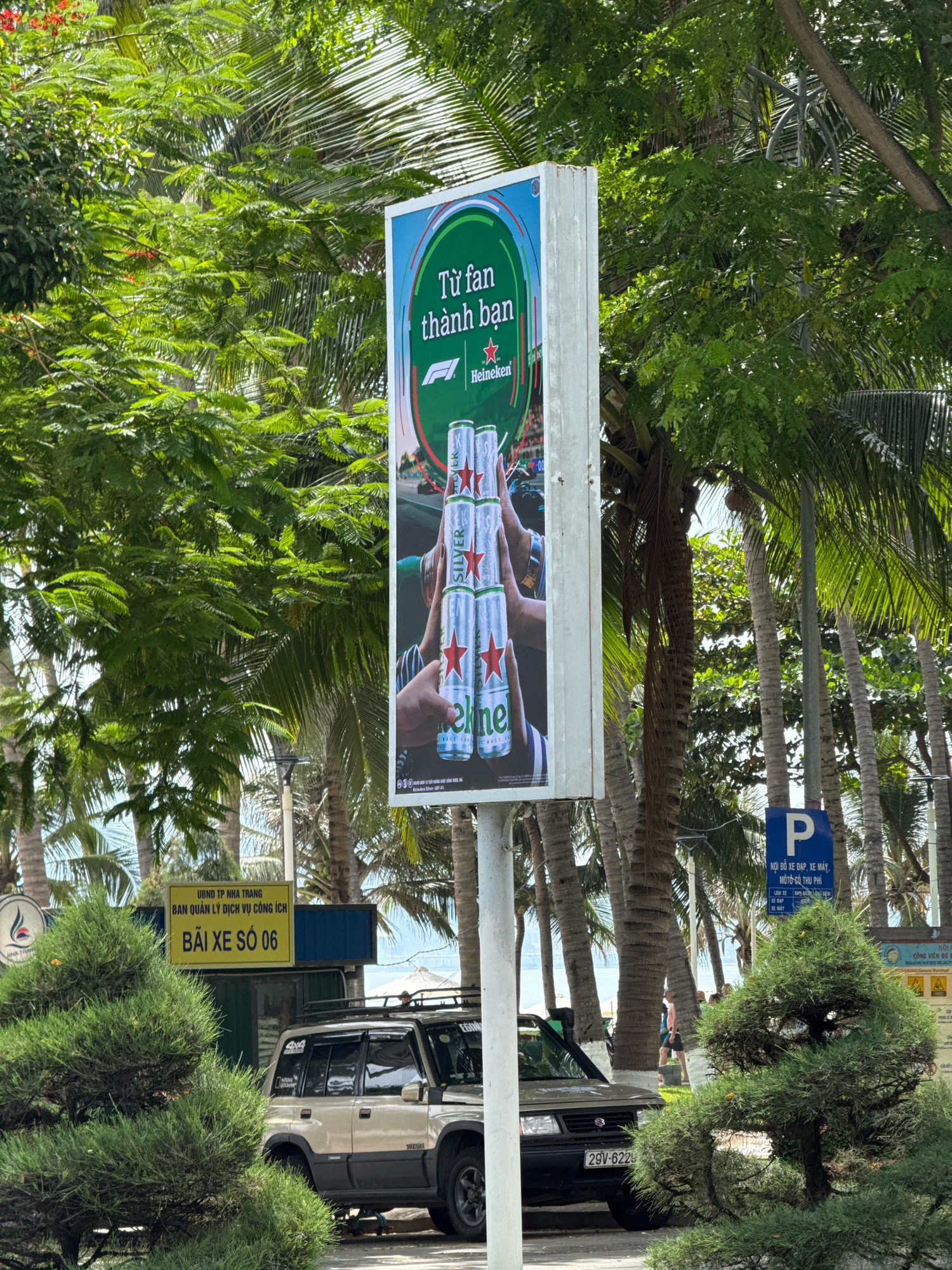 In Nha Trang, Vietnam, a tall, white billboard advertising Heineken beer stands prominently in a tropical environment, surrounded by lush green palm trees. The billboard features images of Heineken cans and the text "Từ fan thành bạn" which translates to "From fan to friend," along with the Heineken logo and an F1 logo, suggesting a connection to motorsports. To the left of the billboard, a yellow sign reads "UBND TP. NHA TRANG BAN QUAN LY DICH VU CONG ICH BAI XE SO 06," indicating a public service parking lot number 06 managed by the Nha Trang City People's Committee. A light-colored SUV with a roof rack is parked in front of this sign. To the right of the billboard, a blue parking sign with a white "P" symbol is visible, along with more text in Vietnamese. The scene is bathed in natural daylight, with sunlight filtering through the dense foliage, creating a bright and vibrant atmosphere.