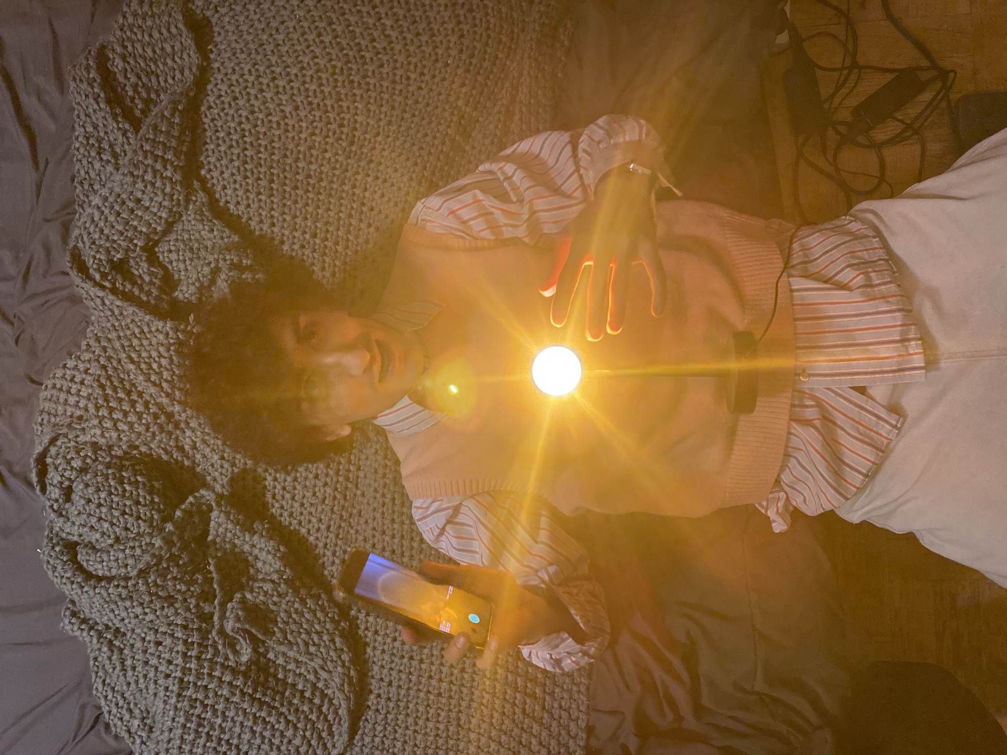 A person is lying on a gray bed with a knitted gray blanket. The person is wearing a striped shirt and a beige vest. They are holding a glowing orb in their hands, and a smartphone in their other hand. There are wires and other items near the bed on the right side of the image. The location is not discernible.