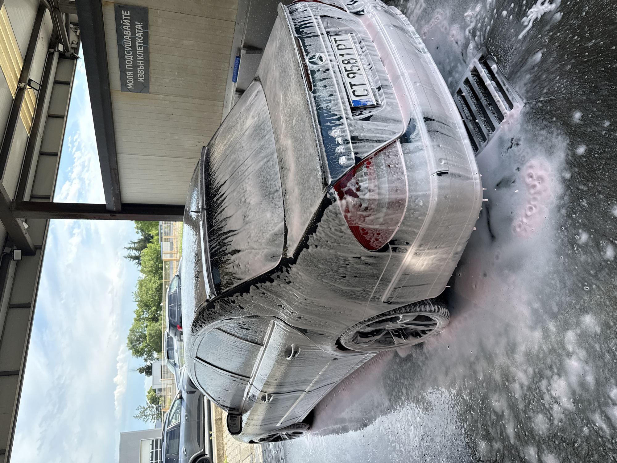 The image depicts a black Mercedes-Benz C320 sedan being washed at a self-service car wash bay in Stara Zagora, Bulgaria, under a partly cloudy daytime sky. The car is extensively covered in thick white and pink-tinged soap suds, with foamy water splashing around its wheels and on the ground, flowing towards a metal drainage grate.
The vehicle's rear right side is most prominent. Visible on the trunk are the Mercedes-Benz emblem and the model designation "C320," partially obscured by foam. The Bulgarian license plate "CT 9581 PT" is clearly displayed, featuring the "BG" country code, and a subtle "MADE IN ITALY" inscription is on its frame.
The setting includes a metal canopy structure overhead, from which a sign with Cyrillic text hangs. The text reads: "МОЛЯ ПОДСУШАВАЙТЕ ИЗВЪН КЛЕТКАТА!", translating to "PLEASE DRY OUTSIDE THE BAY!" Beyond the immediate car wash bay, green trees, other parked cars, and distant buildings are visible, indicating an outdoor environment. The bright natural light suggests it is either morning or afternoon.