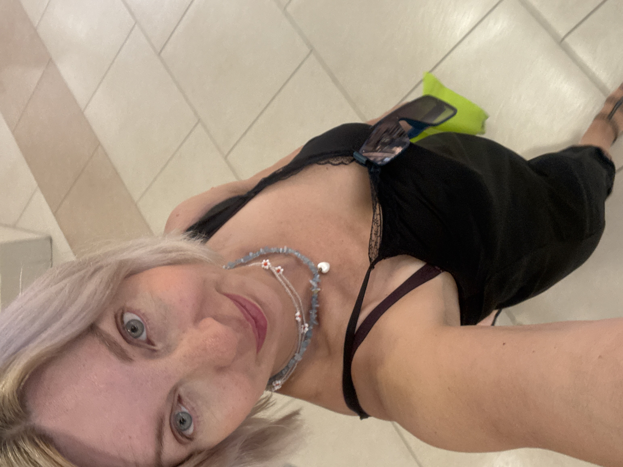 A blonde woman with light blue eyes and fair skin poses for a selfie in an indoor setting with light-colored tiled flooring. She is wearing a black dress with thin straps and a dark bra visible underneath. A beaded necklace with small red and white flowers and a pearl pendant adorns her neck. She has a bright yellow bag and sunglasses hanging from her dress. The woman's hair is short and light blonde, styled in a bob. The background shows large, square beige and white tiles, suggesting a public indoor space. The lighting appears to be overhead. The image is a close-up, focusing on the woman's face and upper body.
