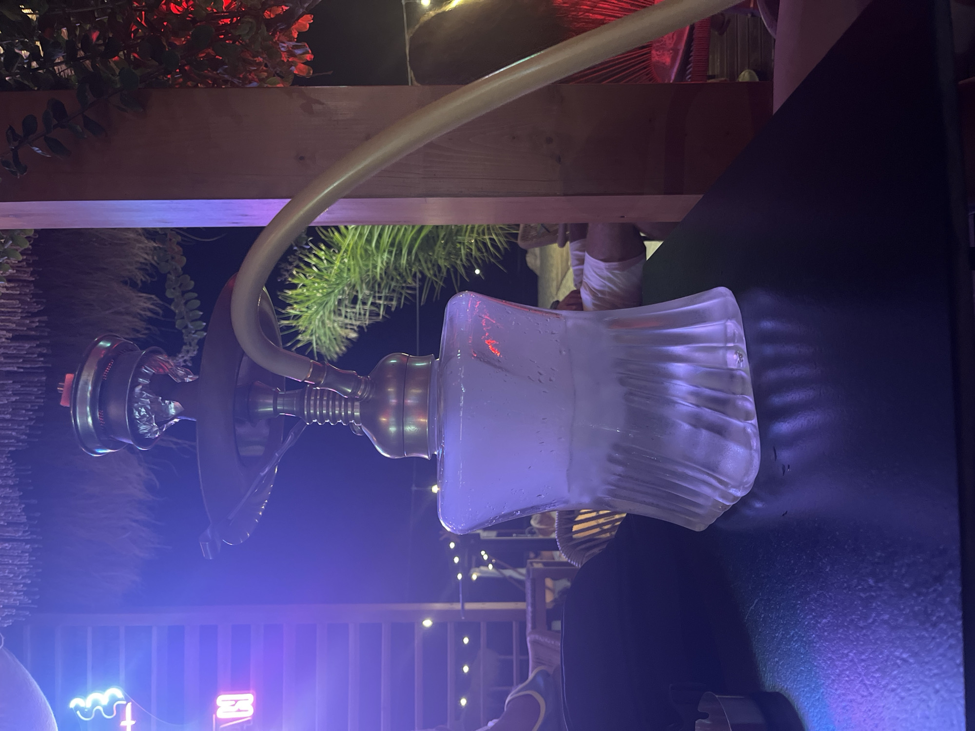 In Bashkia Durrës, Albania, a hookah is displayed on a dark, reflective surface, possibly a table. The hookah features a clear, fluted glass base filled with white vapor, indicative of smoke. Above the base, there is a metallic stem with ribbed detailing, leading to a metallic bowl. A light-colored hose extends from the side of the hookah. The background is dimly lit, with hints of blue and purple ambient lighting, and string lights can be seen in the distance. To the left, neon lights spelling out "N" and a wave-like symbol are visible, suggesting a nighttime establishment. Green foliage and red lights are present in the midground and background. Several out-of-focus figures are partially visible, some seated and others appearing to stand. The overall scene suggests an outdoor or semi-outdoor entertainment venue.