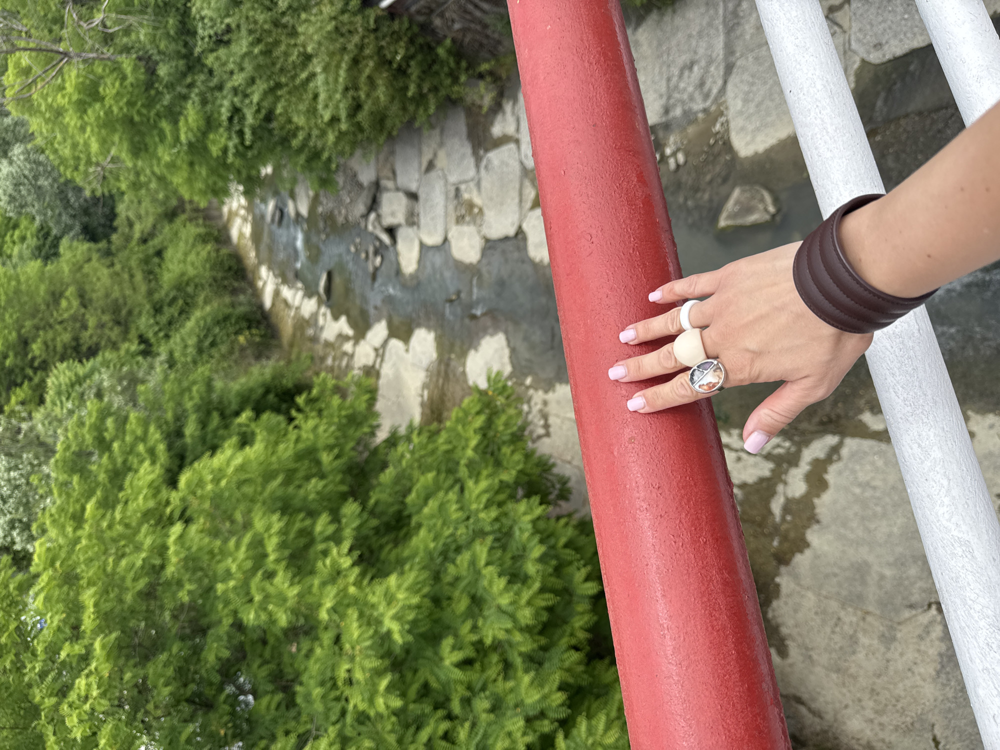 A person's hand with pink fingernails rests on a red railing above a shallow, rocky stream. The person is wearing two rings, one with a circular design and the other a pale beige color, and a dark brown leather cuff bracelet. Lush green trees line the banks of the stream. This scene is likely located in the city of Sochi, Russia, given the presence of a stream and abundant vegetation often found in the region.