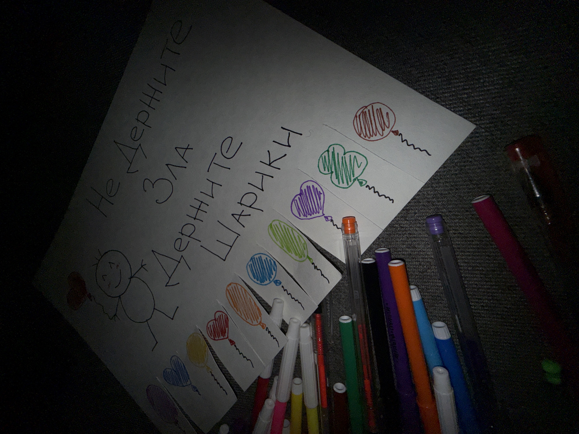 The image shows a piece of paper with a child-like drawing of a stick figure holding a red heart-shaped balloon. To the right of the figure, the paper has been cut into strips, each strip featuring a differently colored balloon. The text "Не Держите зла, Держите Шарики" (Don't hold grudges, hold balloons) is written across the top of the paper. Several colored markers are scattered near the paper. The location, Dmitrov, Russia, is not ascertainable from the given image.