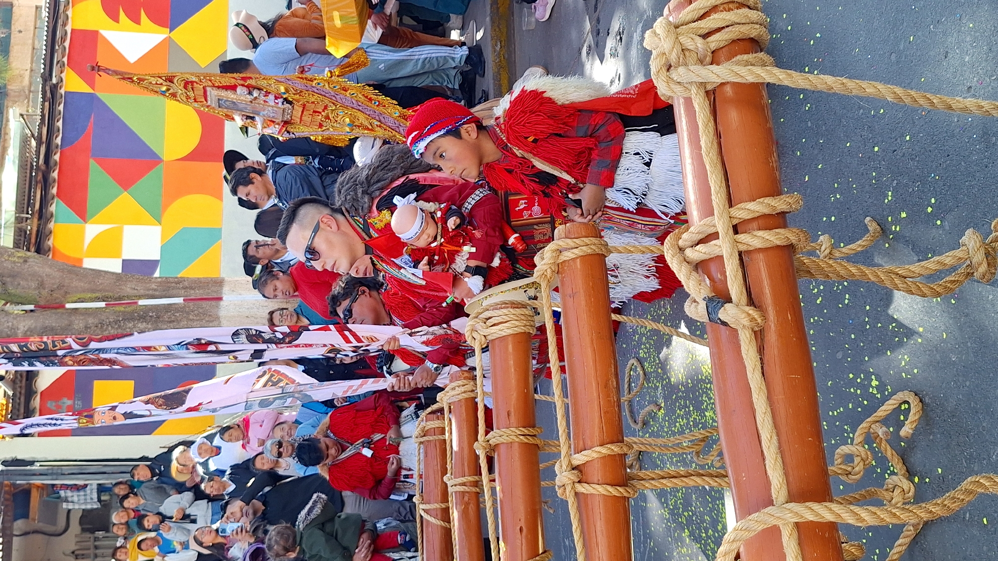 In the city of Wanchaq, Peru, an outdoor procession or parade is underway, indicated by the presence of multiple participants in traditional attire and decorative elements. The foreground features several thick, orange wooden poles supported by ropes, suggesting they are part of a float or carried structure. A young boy in a red and white feathered costume and a red knitted cap looks upwards with clasped hands. To his left, an adult man in similar red attire is holding a doll. Other individuals, dressed in red, white, and patterned clothing, are visible in the background, some holding flags. A vibrant, geometric mural adorns a wall behind the crowd, adding a splash of color to the scene. The ground is littered with confetti.