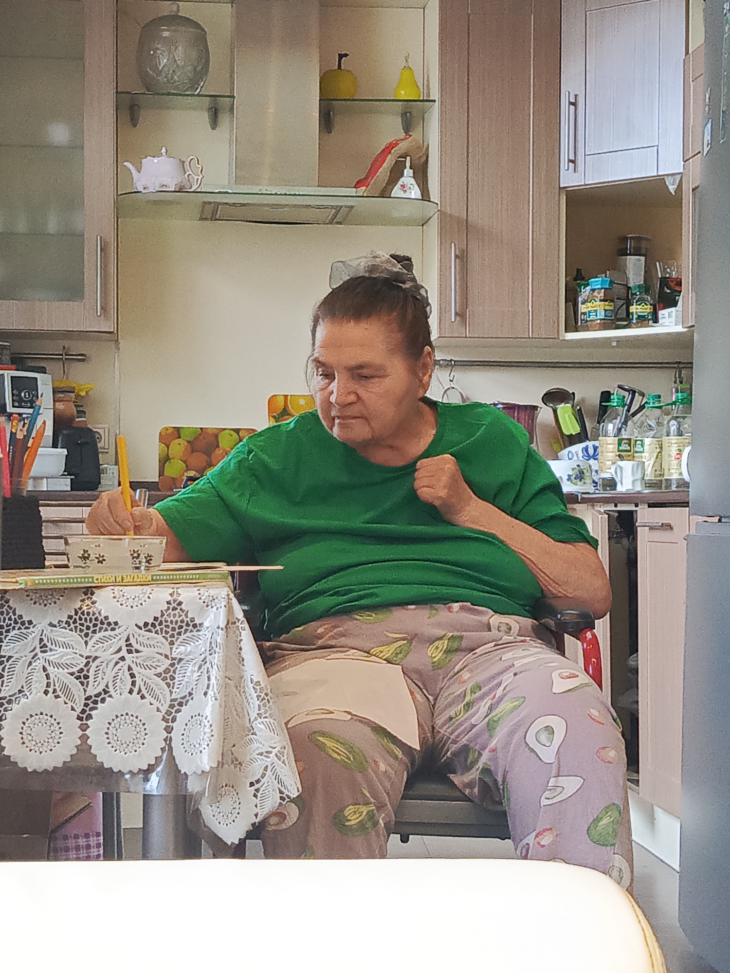 A full shot shows an elderly woman sitting in a wheelchair at a table in a kitchen. She is wearing a green t-shirt and patterned pants. Her hair is pulled back. She is holding a yellow object, possibly a pen or crayon, and appears to be drawing or writing in a bowl on the table. The table is covered with a white lace tablecloth. In the background, kitchen cabinets are visible, some of which contain jars and bottles. A shelf above the table holds decorative items, including a teapot and a yellow apple. The overall impression is of a domestic scene, likely within a residential setting in Saint Petersburg, Russia, where the woman is engaged in a quiet activity.