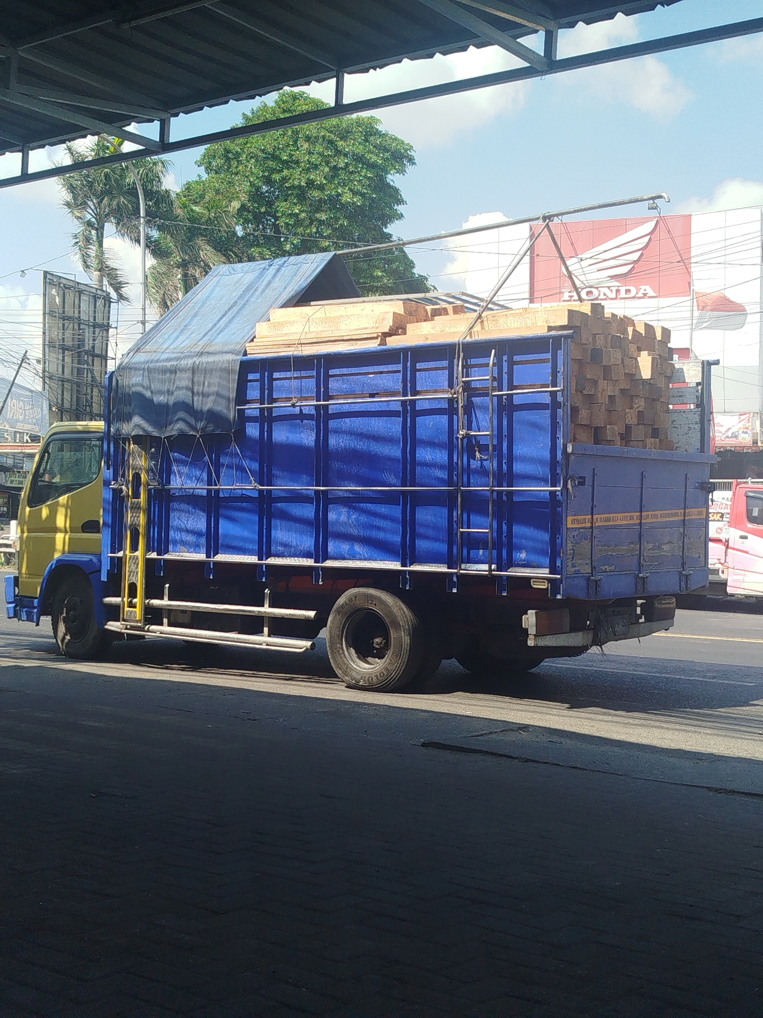 A yellow and blue flatbed truck, laden with wooden planks covered by a blue tarp, drives down a paved road in Sukomulyo, Indonesia. The truck has a ladder attached to its side, and the wooden planks are stacked high in the bed. In the background, a large red and white Honda logo is visible on a building, and trees and a cloudy sky can be seen. The image is taken from a low angle, under an awning.