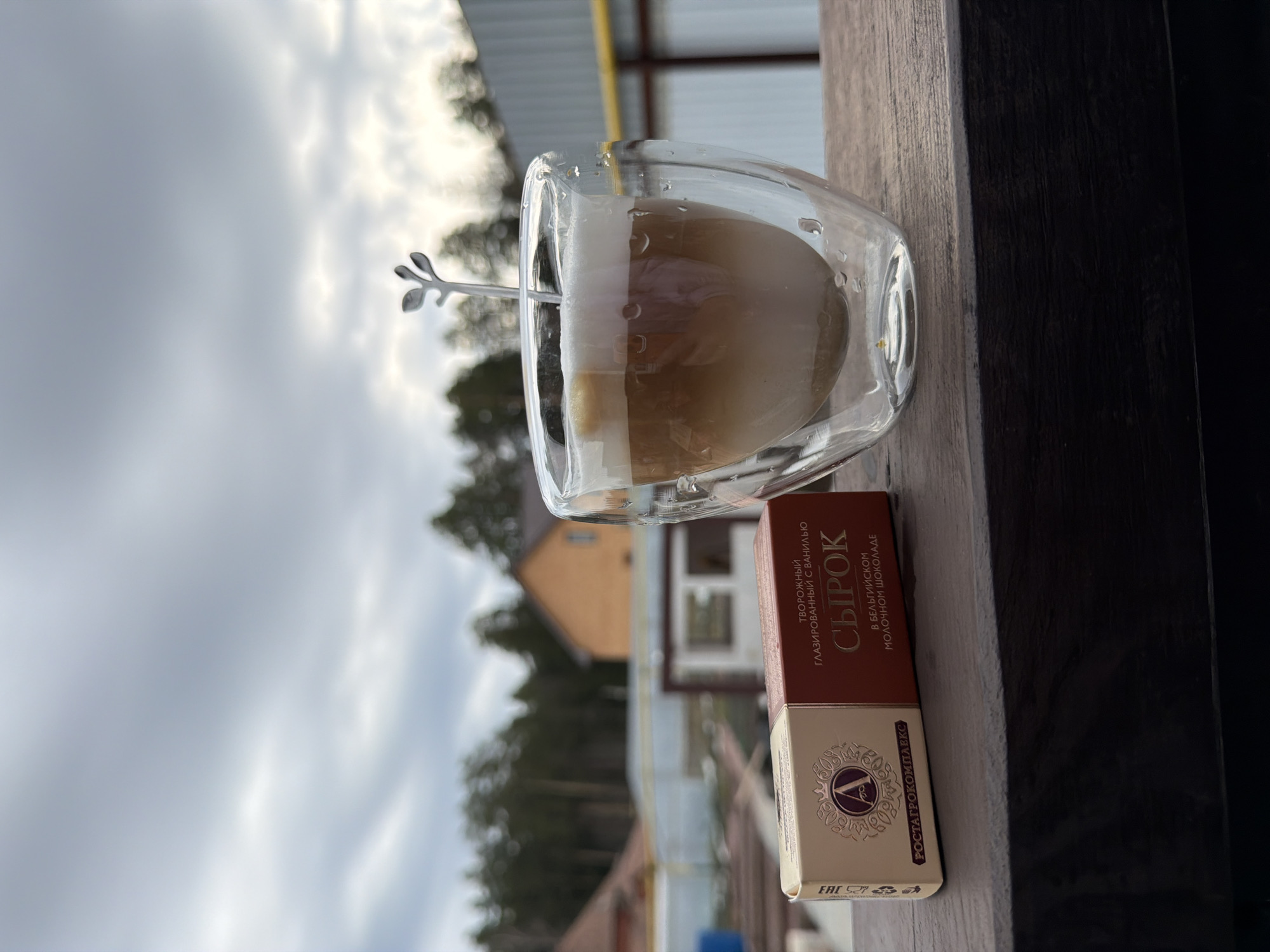 The image shows a glass of coffee with a spoon in it and a box of "Syrok" glazed curd cheese with vanilla in Belgian milk chocolate on a wooden surface. In the background, there's a house surrounded by trees and an overcast sky. The photo appears to be taken outdoors, possibly on a balcony or patio.
The location is Chaikovsky, Russia.
