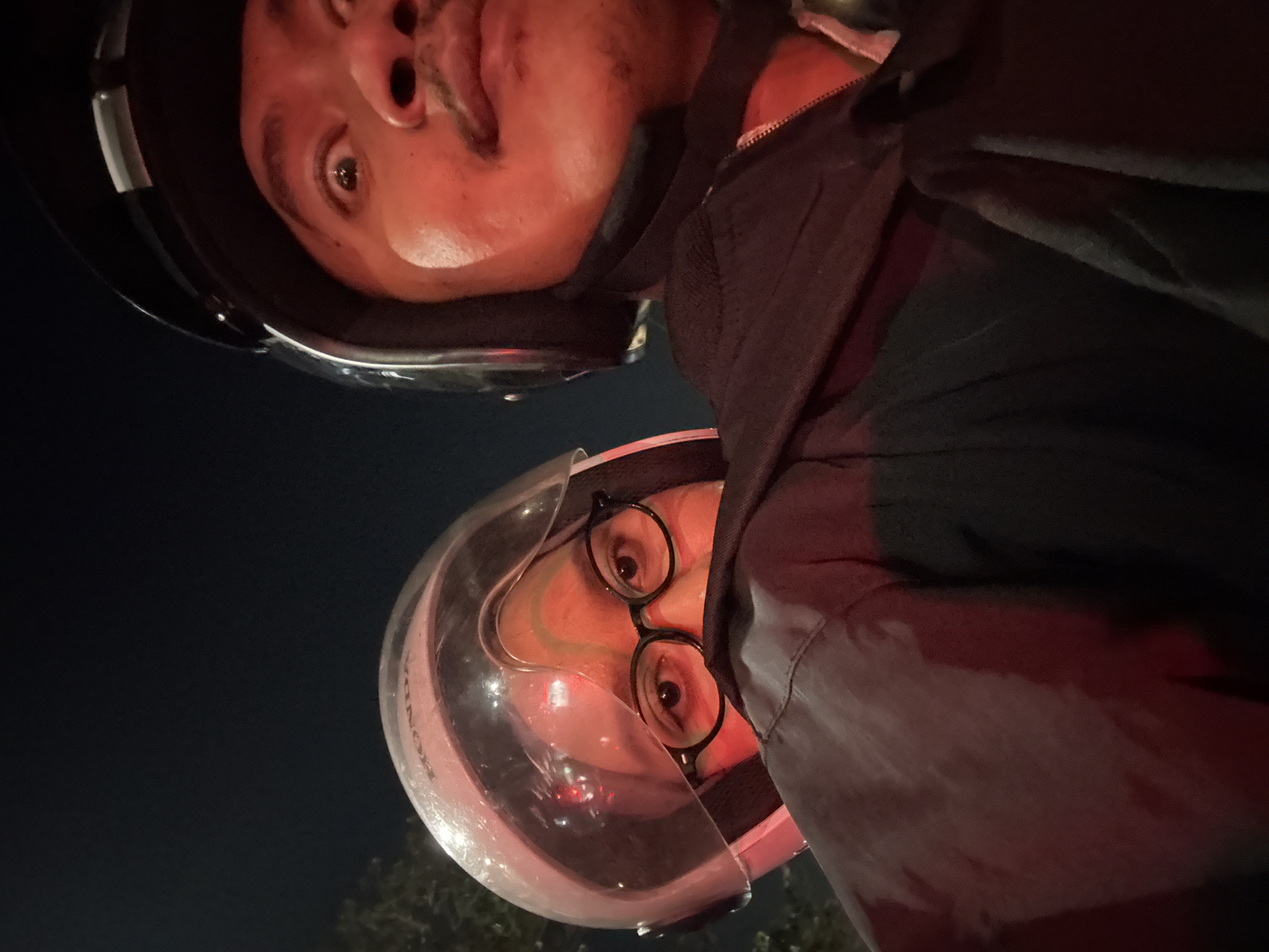 In this close-up selfie taken at night, two individuals are positioned on a motorcycle. The person in the foreground, on the right, wears a black helmet and appears to be looking at the camera with wide eyes and a surprised expression. Their face is illuminated by a red light, highlighting their mustache and skin. To the left and slightly behind, a second person wears a light pink helmet with a clear visor. They are partially obscured by the first person's shoulder and jacket but their eyes, visible behind glasses, are looking forward. The background is dark and indistinct, suggesting an urban night environment, possibly along a street in West Jakarta, Indonesia, given the presence of streetlights and the overall ambiance of nighttime city travel. The clothing suggests they are dressed for riding.