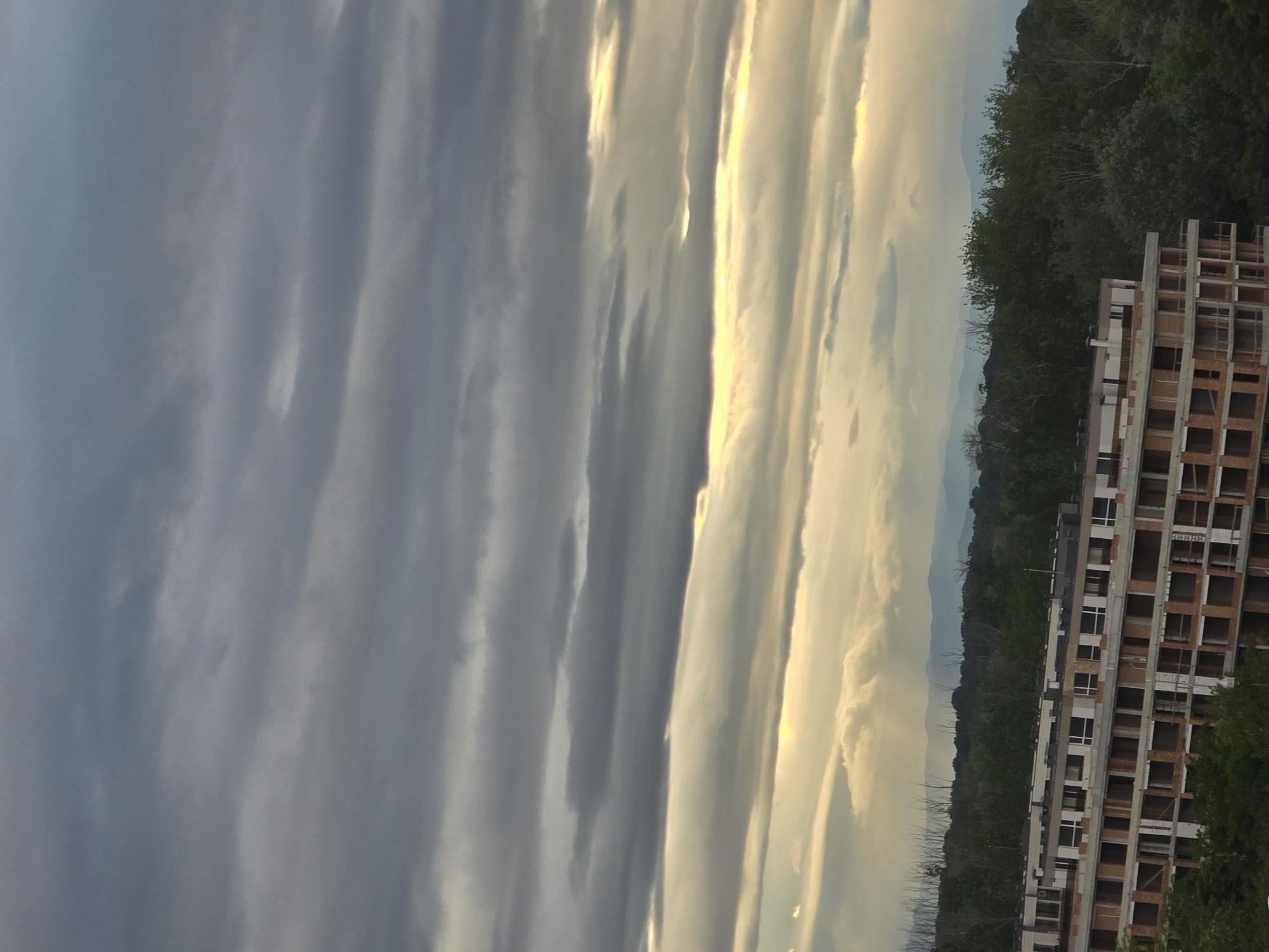 The image shows an unfinished building under a cloudy sky. The building is multi-storied and constructed of brick and concrete. To the left of the building is a dense green forest. In the distance, hazy mountains can be seen. The sky is filled with long, striated clouds, transitioning from dark grey to light yellow. The image appears to be taken at dusk or dawn. The location is Unknown, Unknown.