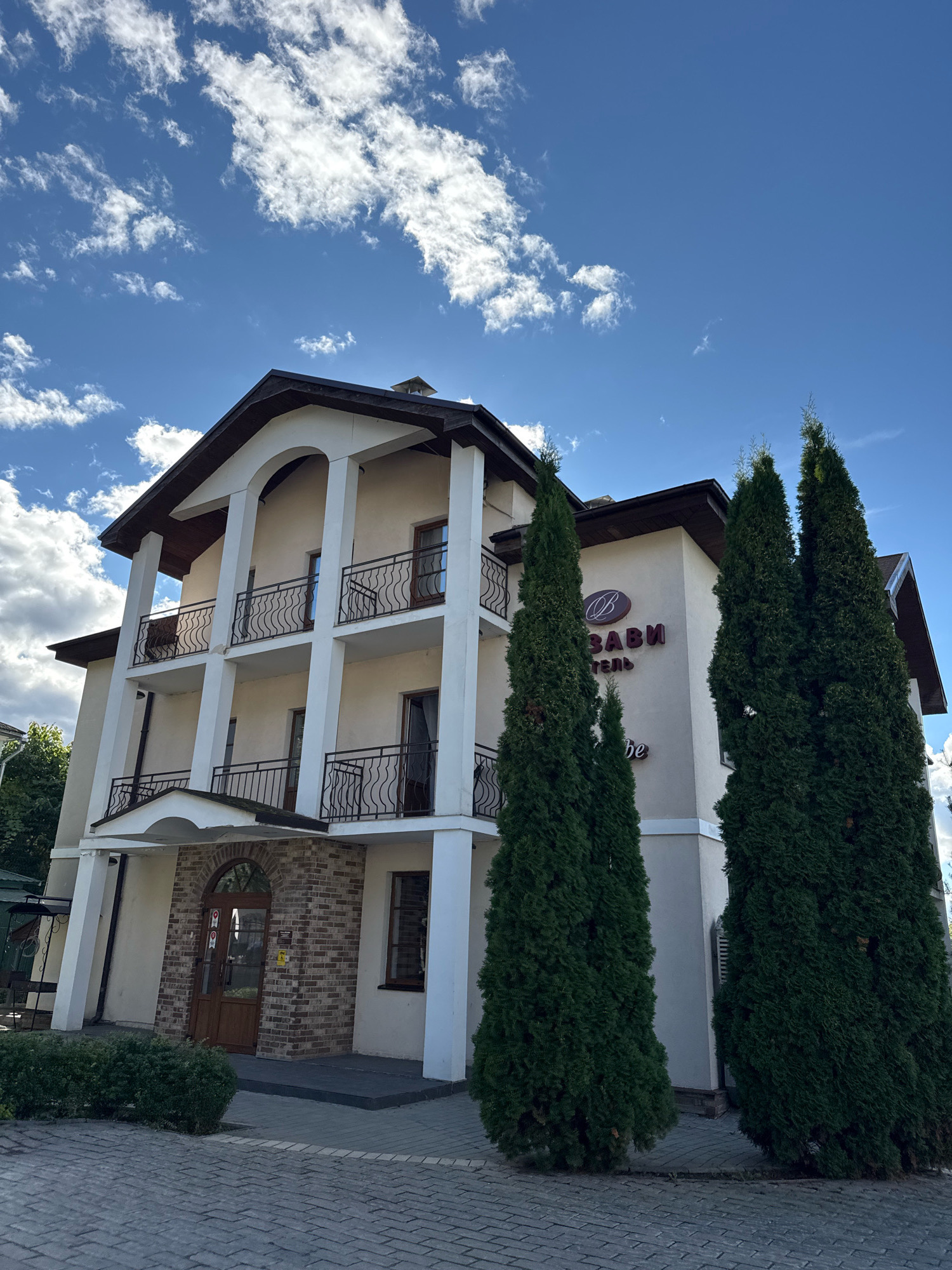 The image shows a three-story hotel building in the city of Staraya Russa, Russia. The building has a light-colored facade with white pillars supporting balconies on each level. The balconies have black wrought-iron railings. The entrance is beneath an arched brick facade. Two tall, dark green evergreen trees stand in front of the building. The sky above is blue with scattered white clouds. Text on the building indicates it is a hotel.