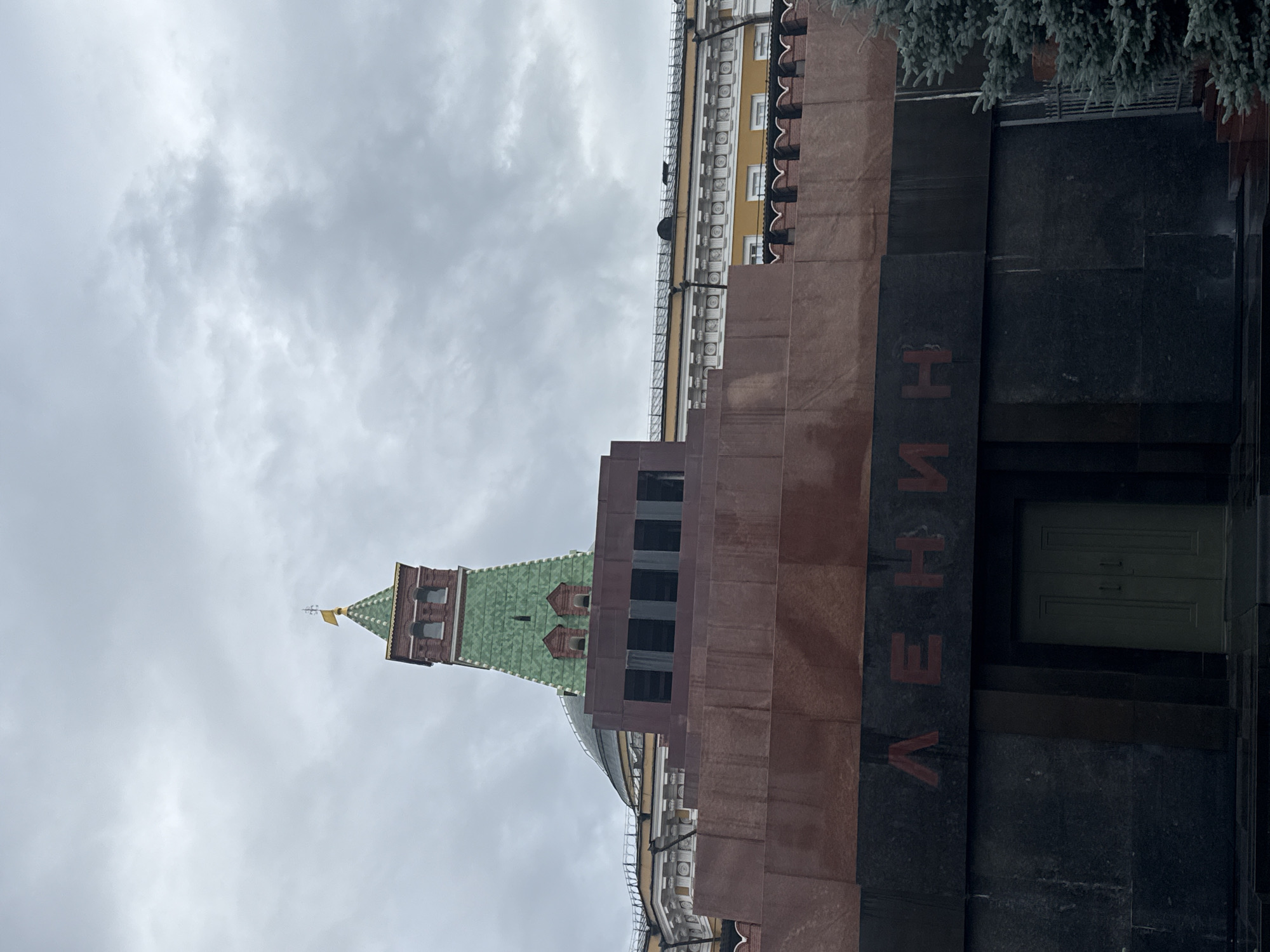 This is a close-up, vertically oriented shot of a monument with a red-brick tower and a green, multi-faceted roof, topped with a small yellow flag. The tower is part of a larger structure made of dark stone and reddish-brown granite. Red lettering is visible on the dark stone, spelling out "ЛЕНИН" (Lenin in Russian). The background is a cloudy, overcast sky. The overall mood is somber and overcast, fitting for a monument. The lighting suggests it could be daytime, but the clouds obscure any direct sunlight. The architecture points to a historical or governmental location, likely in Russia given the Cyrillic text. There are no people visible in the image.
