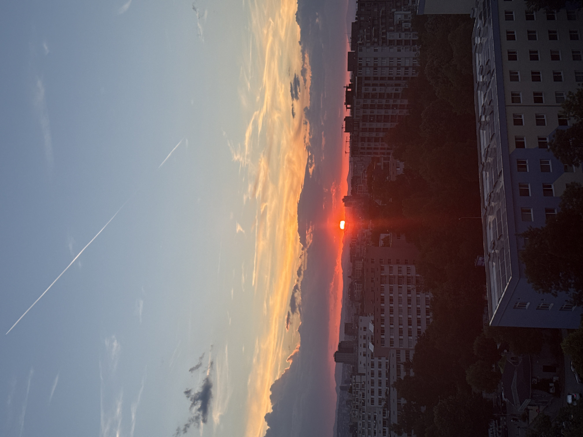 This image depicts a sunset over the city of Sofia, Bulgaria, likely taken from a high vantage point. The sky is a dramatic canvas of colors, transitioning from a pale blue at the top to vibrant oranges and reds closer to the horizon, illuminated by the setting sun. Dark, heavy clouds are scattered across the lower half of the sky, contrasting with wispy, lighter clouds higher up. Two faint white contrails from aircraft are visible in the upper sky.
Below the sky, the urban landscape of Sofia unfolds. A dense collection of apartment buildings dominates the midground. Many of these buildings are tall, multi-story structures with a uniform architectural style, characteristic of Soviet-era housing blocks. The buildings are packed closely together, interspersed with large areas of dark green trees, suggesting residential neighborhoods with significant tree cover.
In the foreground, the top of a lower-rise building is visible, featuring a light blue and yellow facade and a roof covered with what appear to be solar panels. A dark, shadowed area below this building suggests more trees and potentially some smaller structures or roads. The overall impression is of a dense urban environment bathed in the warm, fading light of sunset. There are no discernible people or active interactions in the scene.