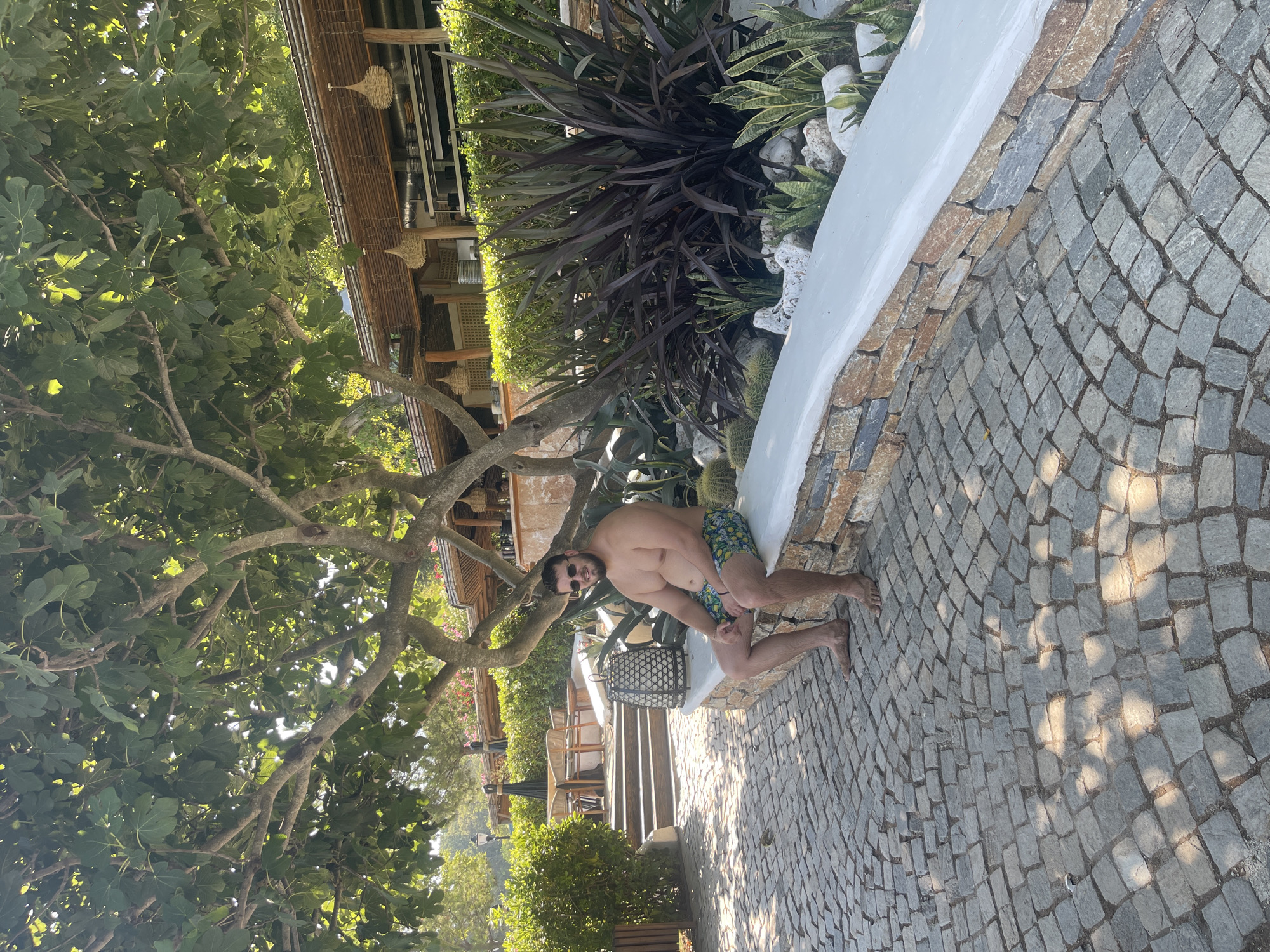 A man with a beard and sunglasses is sitting on a white stone ledge in an outdoor setting. He is wearing patterned swim trunks and is shirtless. He is holding a small object in his right hand. The ledge is made of stacked stone on the lower portion and a smooth white surface on top. In front of the ledge, a cobblestone path is visible, with sunlight and shadows creating a dappled effect. Behind the man, there is lush greenery, a tree with large leaves overhead, and a building with a thatched roof structure. In the background, there are outdoor dining tables and chairs. The overall scene suggests a warm, sunny day, likely in a resort or a restaurant with an outdoor area. The location is identified as Chernomorets, Bulgaria.