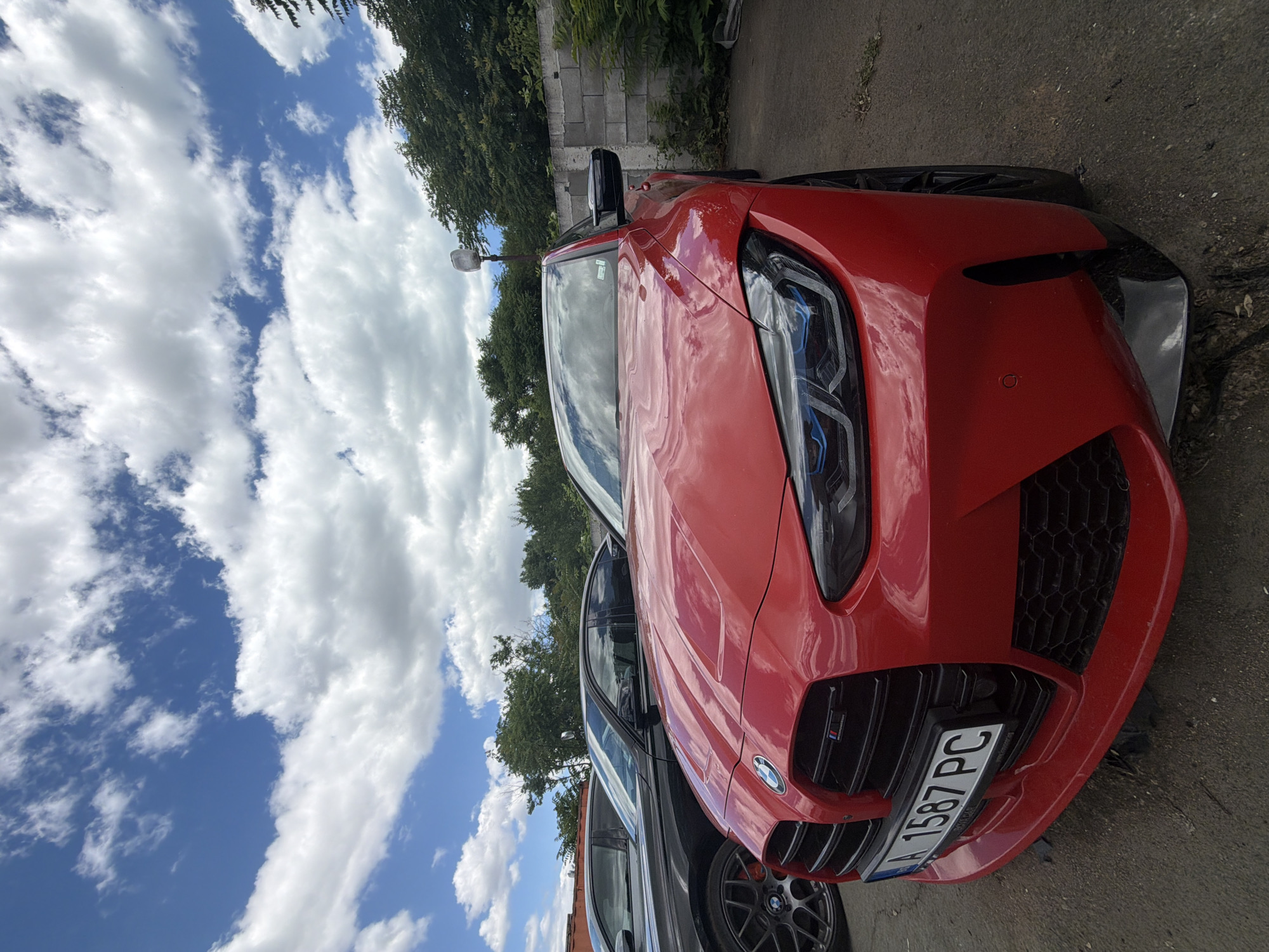 A low-angle, close-up shot captures the front of a red BMW M4 parked outdoors. The car's distinctive kidney grille and aggressive headlights with blue accents are prominently displayed. A license plate reading "A 1587 PC" is attached to the front bumper. To the left, the wheel and fender of a black car are visible. The background consists of a weathered concrete wall, lush green trees, and a bright blue sky filled with dramatic white clouds. The overall setting appears to be a parking area or lot in a semi-industrial or urban environment, potentially in Burgas, Bulgaria, given the license plate format.