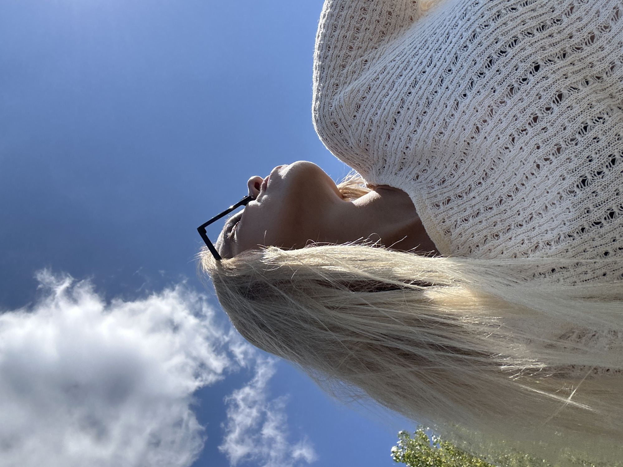 A close-up, low-angle outdoor shot features a woman from the chest up, looking skyward. She has long, blonde hair and is wearing a cream-colored, open-knit sweater and black-framed glasses. The background consists of a bright blue sky with a few scattered white clouds. A portion of a green tree is visible on the far left. The lighting suggests it is daytime. The image is set in Gornyi Erik, Russia.