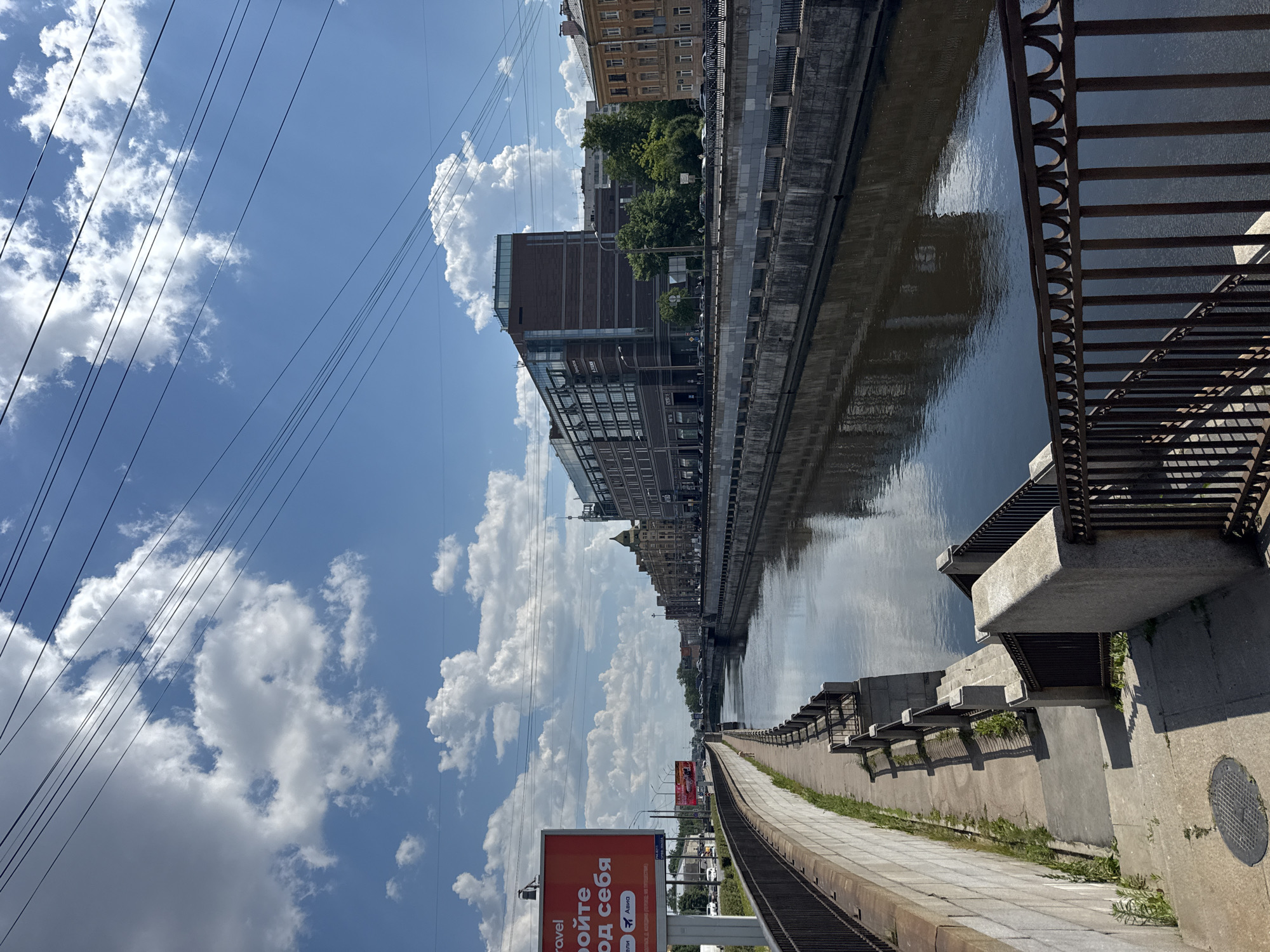 The image captures a vibrant city scene alongside a wide canal or river, observed on a clear, sunny day. The sky is bright blue, dotted with fluffy white cumulus clouds, and crisscrossed by numerous overhead power lines.
Along the left bank of the waterway, a wide concrete embankment features a pedestrian walkway that extends into the distance. A dark, ornate metal railing runs alongside the walkway, while concrete structures with smaller railings lead down towards the water at intervals. Some weeds grow from cracks in the concrete. The canal's surface is dark but reflects the sky and distant buildings, creating shimmering patterns.
On the far bank, a variety of buildings line the waterfront. These include older, ornate multi-story structures and a more modern building with extensive glass windows, all under the bright sunlight. Trees are visible interspersed among the buildings.
In the foreground on the left, a large, prominent orange billboard stands tall. The primary text on the billboard is in Cyrillic, reading "РОЙТЕ ОД СЕБЯ" (likely part of a phrase meaning "find for yourself" or "discover for yourself"). Below this, smaller text includes "travel" and "АВИА" (AVIA, referring to air travel), along with a visible logo resembling "S7", indicating an advertisement for air travel or a related service.
No people are visible in the scene, which appears peaceful and still. The presence of Cyrillic text strongly suggests the location is in a Russian-speaking country.
