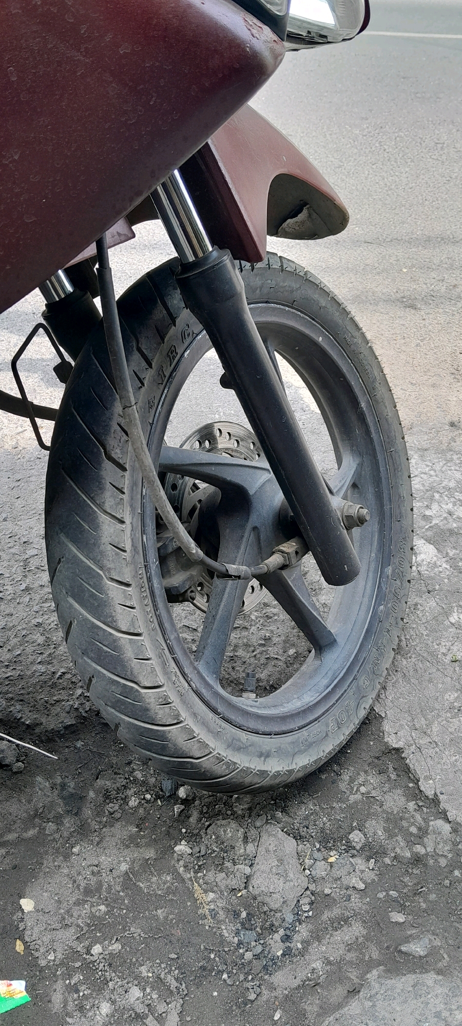 The image shows a close-up of a motorcycle's front wheel and suspension, resting on a paved surface with some debris. The motorcycle's bodywork is a dark maroon color and appears to be made of plastic. The front fork is black, with silver-colored suspension components visible. The wheel is black with a tire that has visible tread markings and text indicating it is "MADE IN INDONESIA" and possibly "IRC". A brake disc is visible behind the wheel hub. The ground is a mixture of cracked asphalt and loose gravel, dirt, and small stones. The scene suggests an outdoor urban or semi-urban environment, possibly in the city of Pasuruan, Indonesia, given the tire's origin. There are no people or any visible actions or interactions taking place. The lighting suggests it is daytime.
